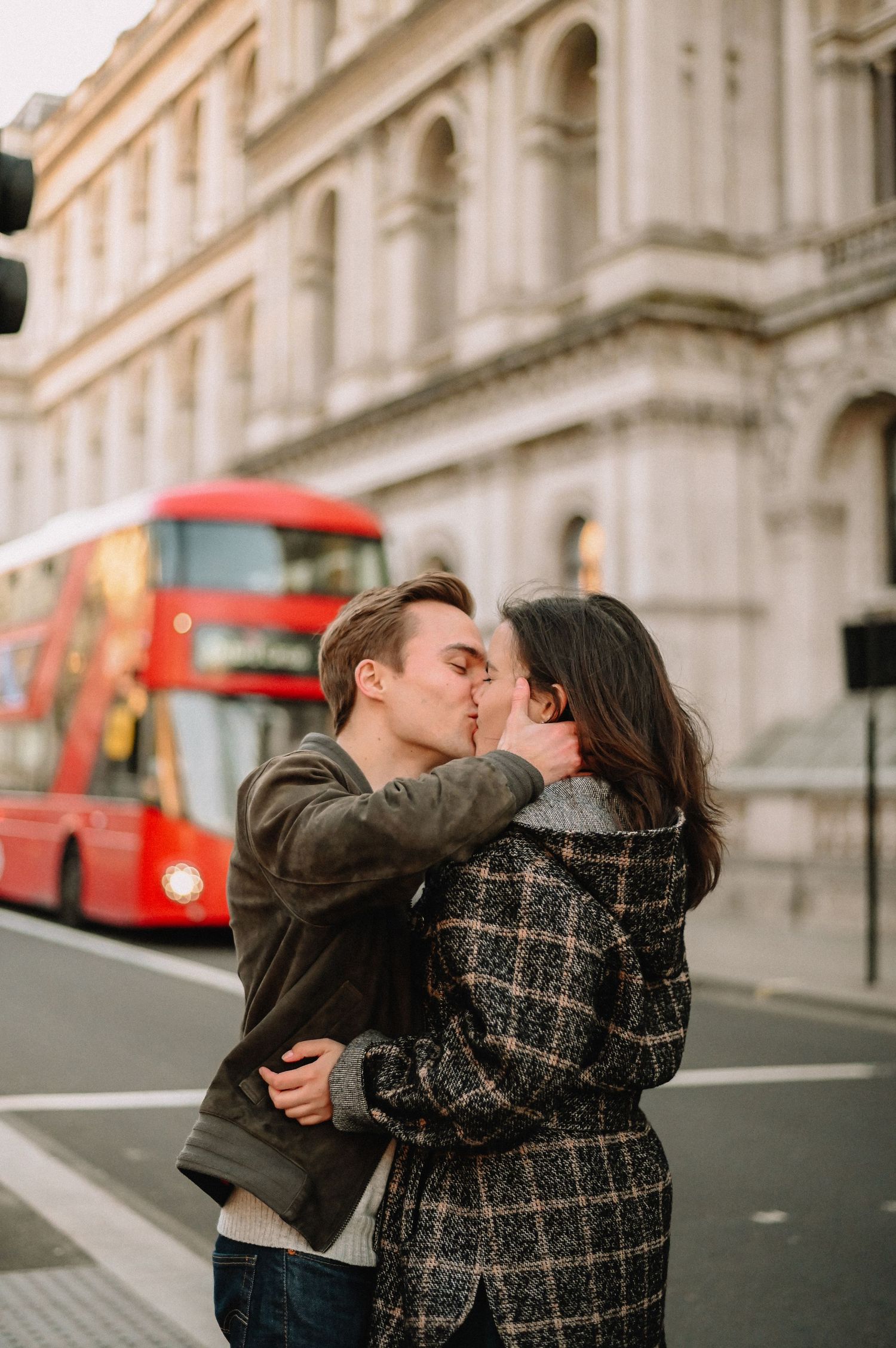 Couple kissing in front of a red double-decker bus on a London street with historic architecture in the background.