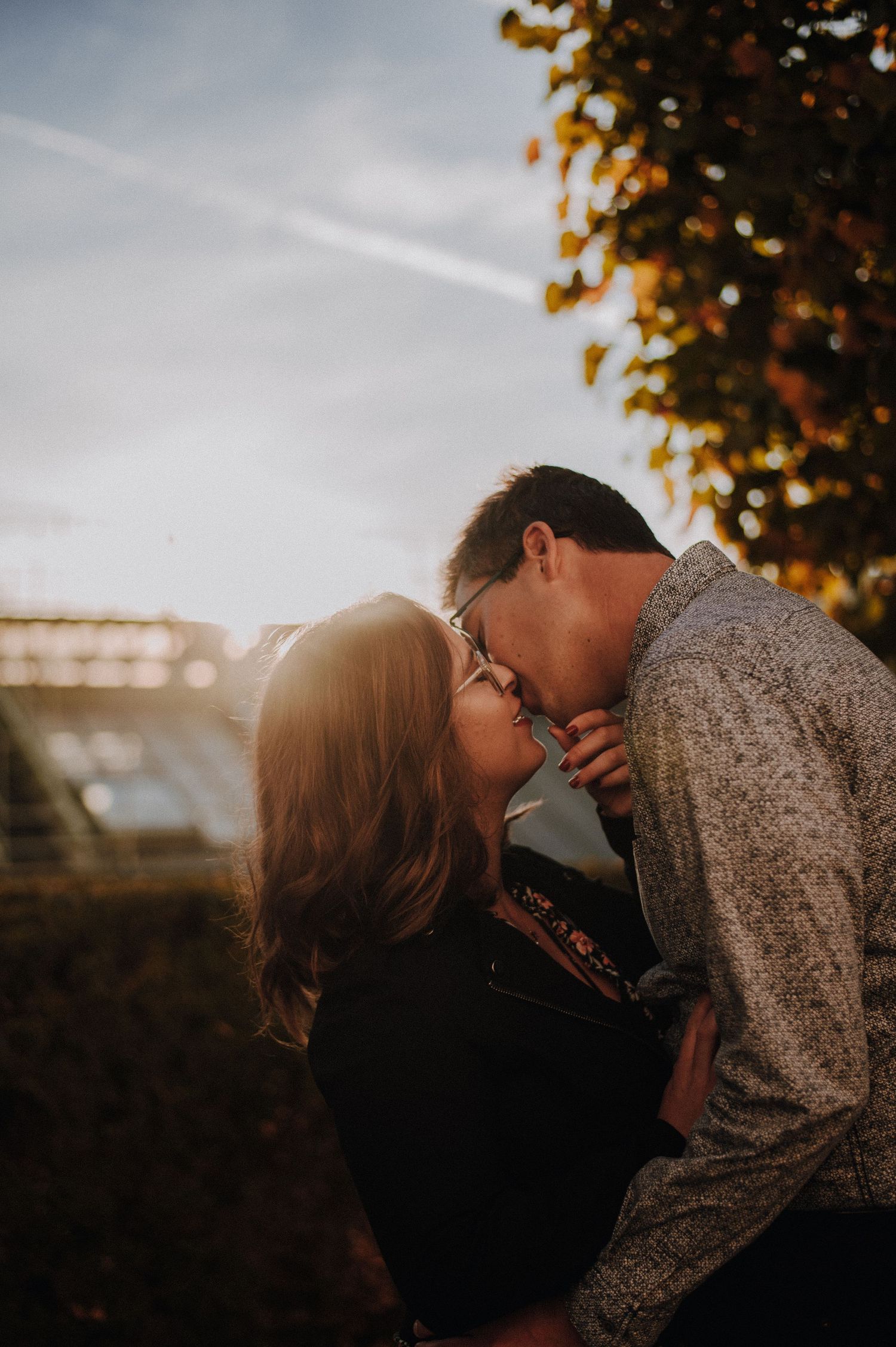 Couple sharing a romantic kiss outdoors during golden hour with autumn trees and warm sunlight in the background.