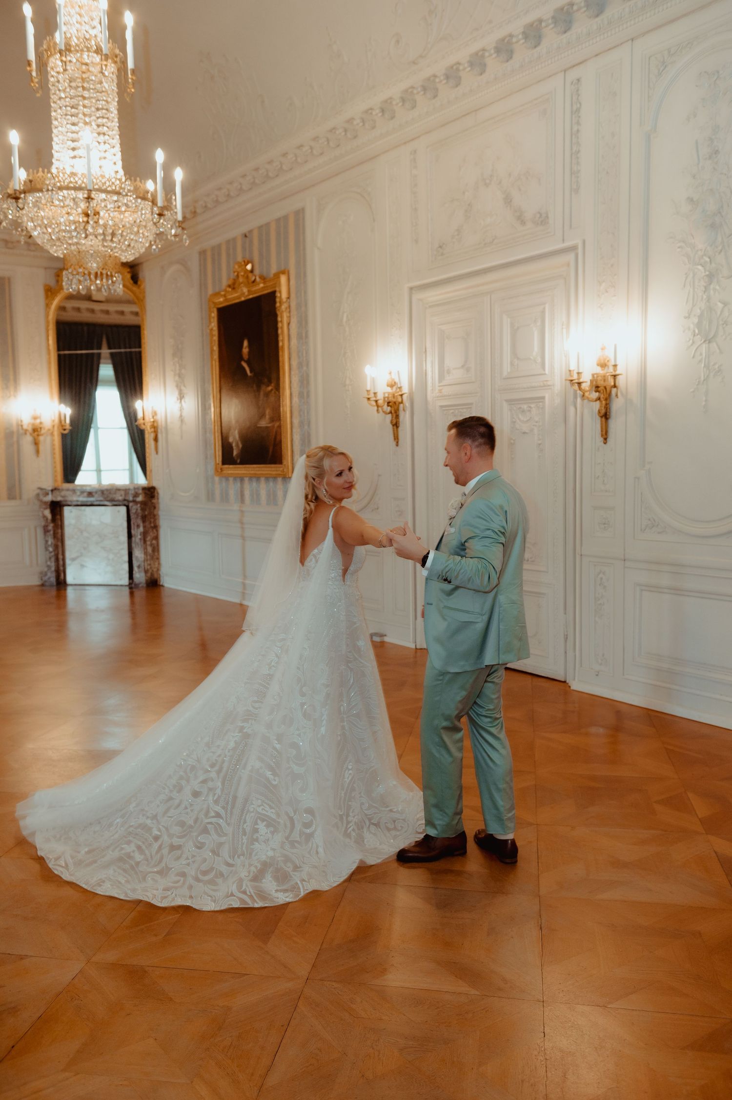 Bride and groom share first dance in elegant ballroom with crystal chandelier and ornate white walls.
