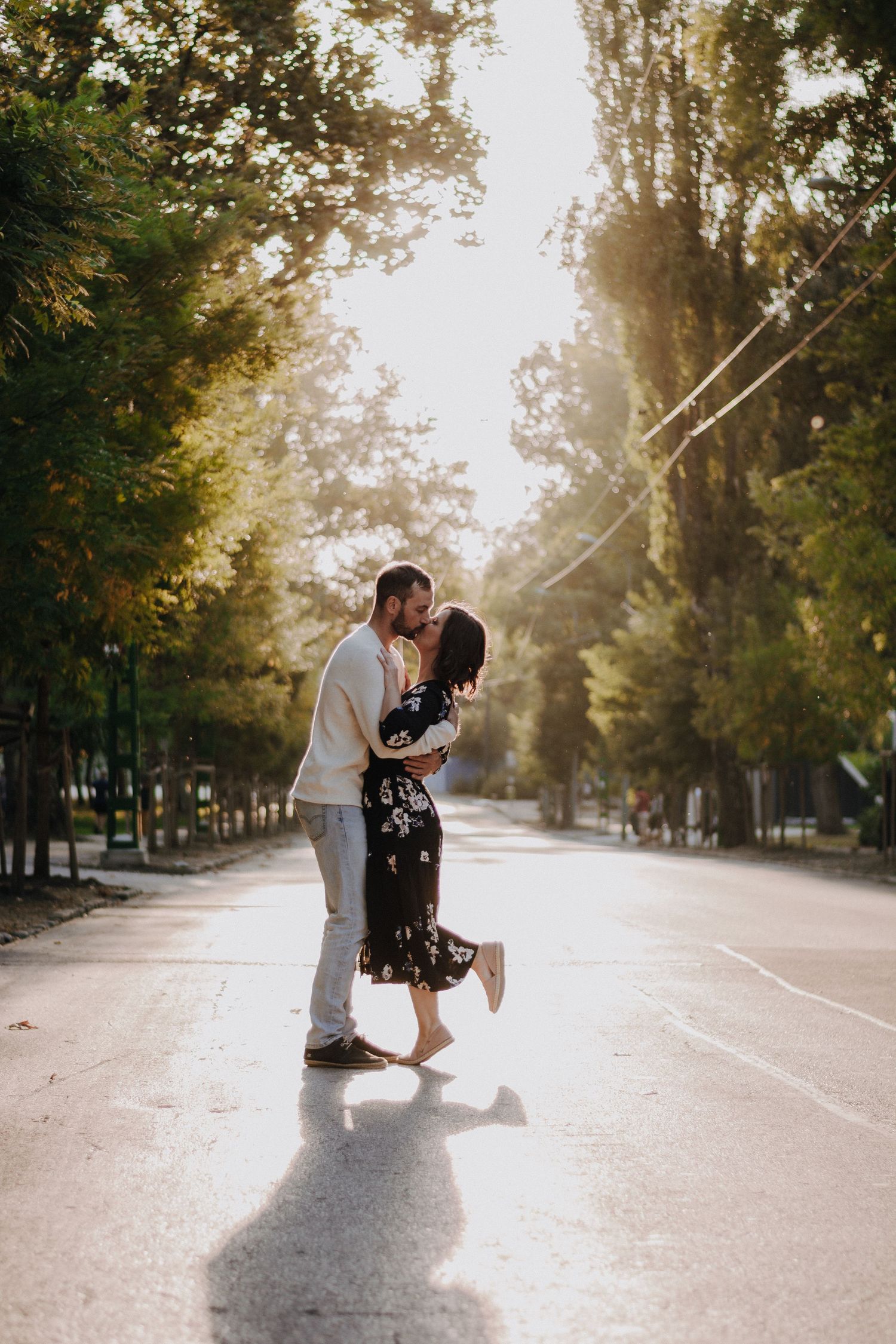 Couple sharing a romantic kiss on a sunlit tree-lined road, woman in floral dress lifting her foot.