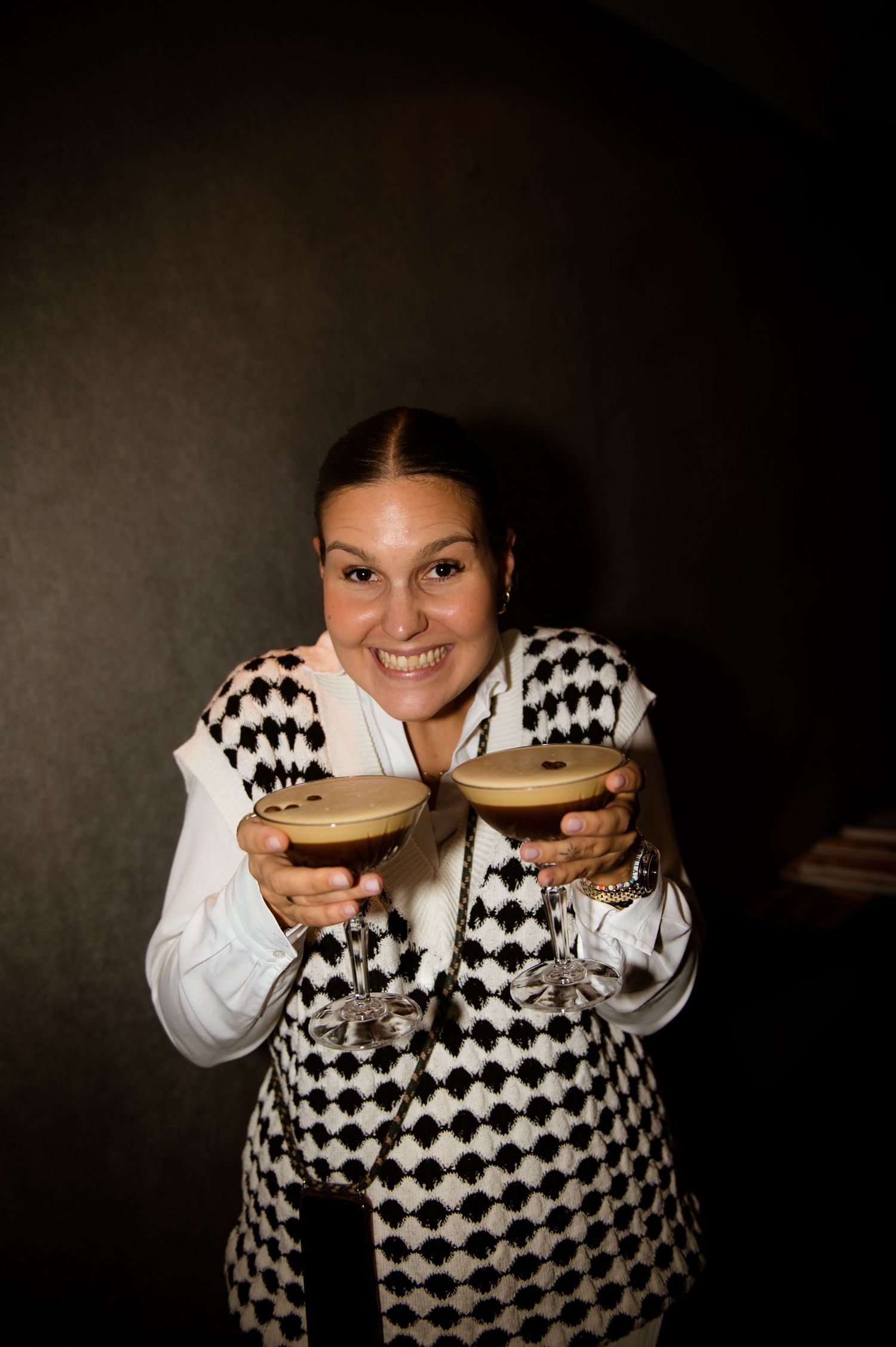 Smiling woman holding two espresso martini cocktails wearing a black and white harlequin vest in a dark bar setting.