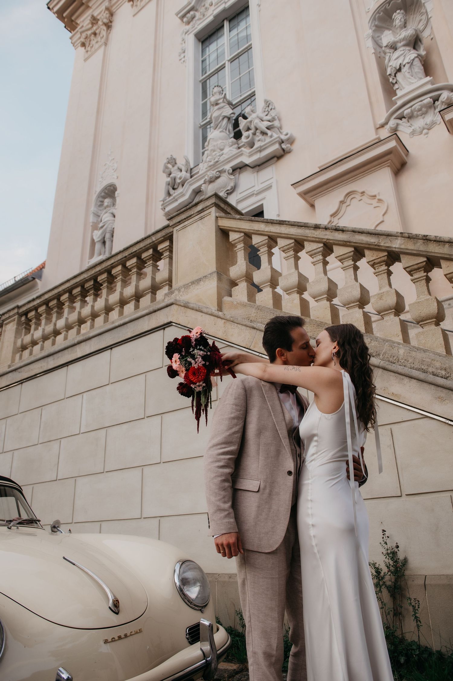 Bride and groom kissing outside ornate building with vintage cream Porsche, bride holding red bouquet in white gown.