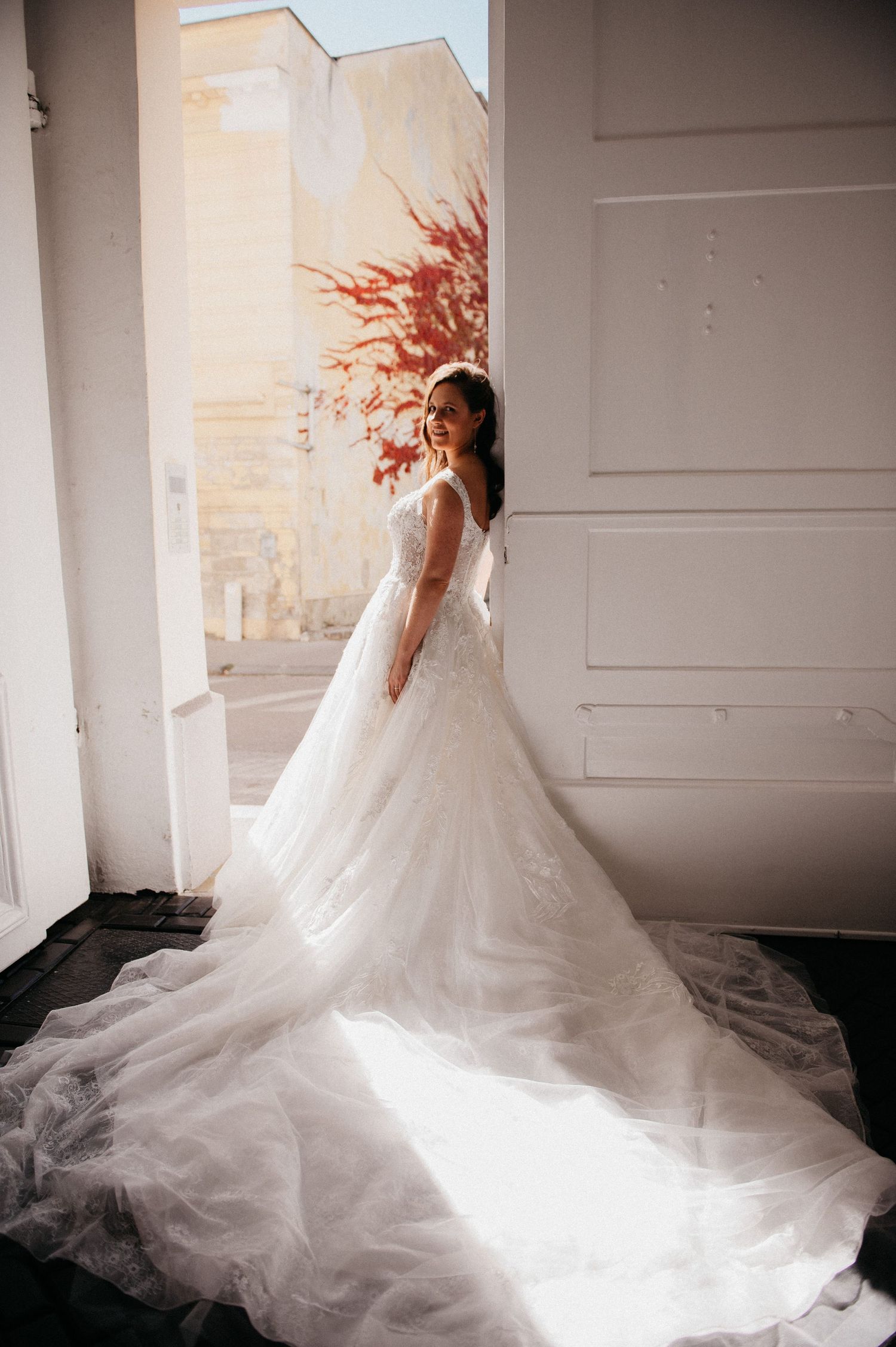 Bride in stunning ball gown wedding dress with long tulle train standing in sunlit doorway.