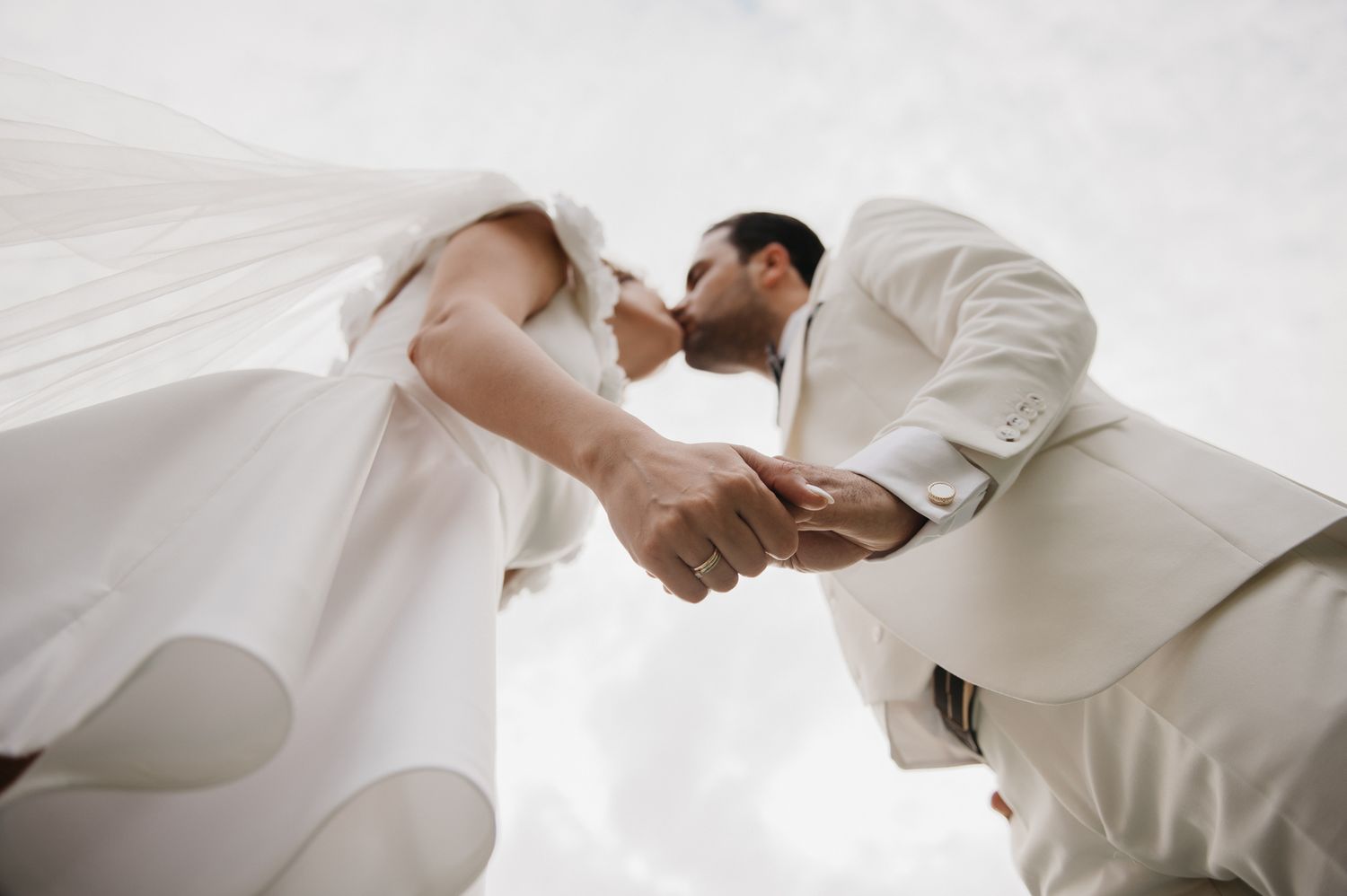 Low-angle view of bride and groom kissing and holding hands against a bright white sky.