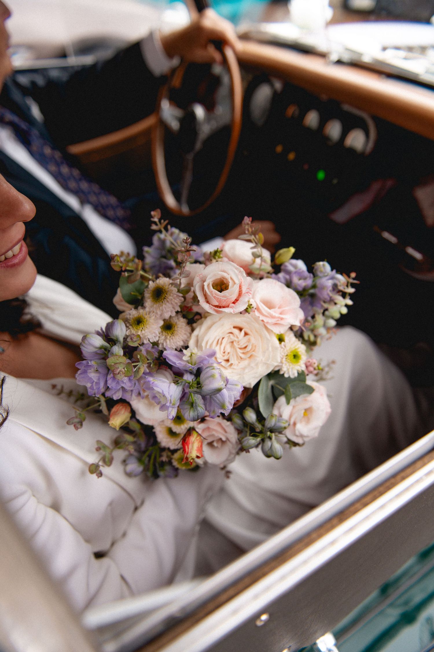 Bride and groom sitting in a vintage car, bride holding a lush pastel bouquet of pink and lavender flowers.