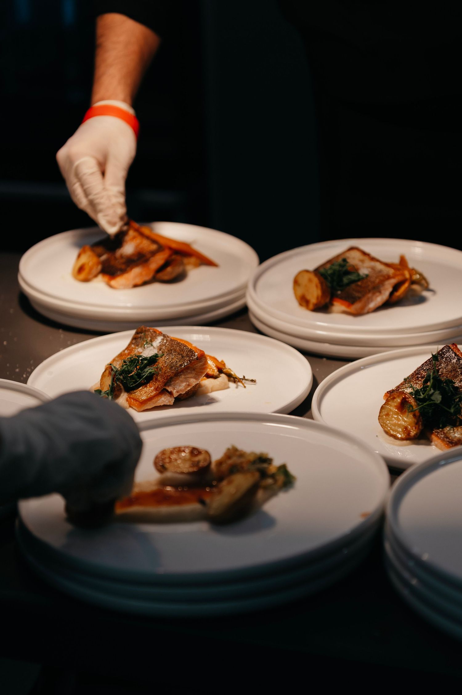 Chefs in gloves carefully plate gourmet fish dishes with vegetables on white plates in a professional kitchen setting.