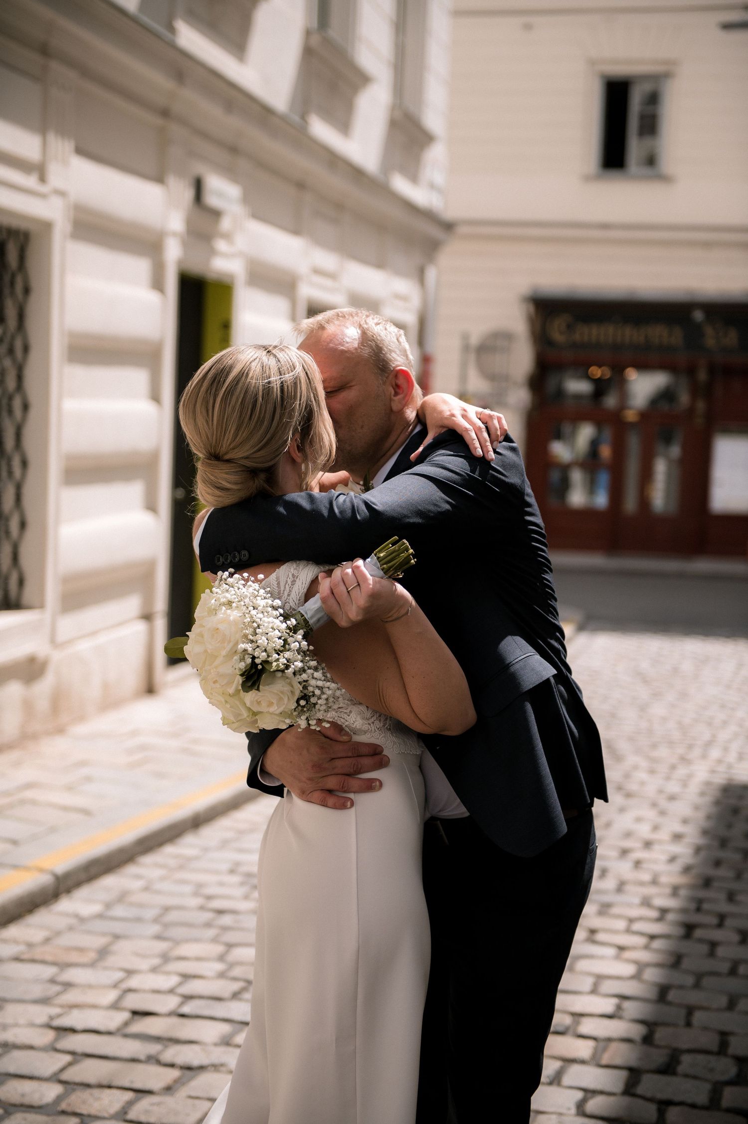 A newlywed couple shares a romantic embrace on a cobblestone street, the bride holding a white bouquet in a elegant dress.