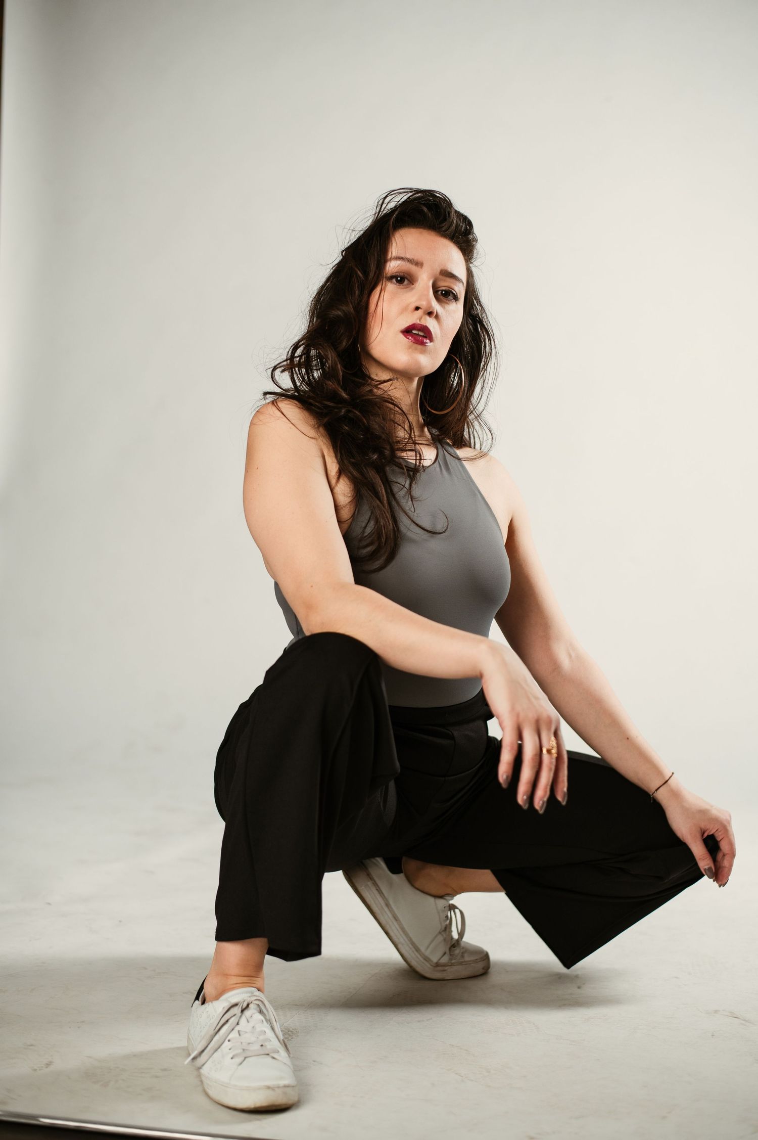Brunette woman in gray crop top and black trousers crouches confidently in white studio setting.