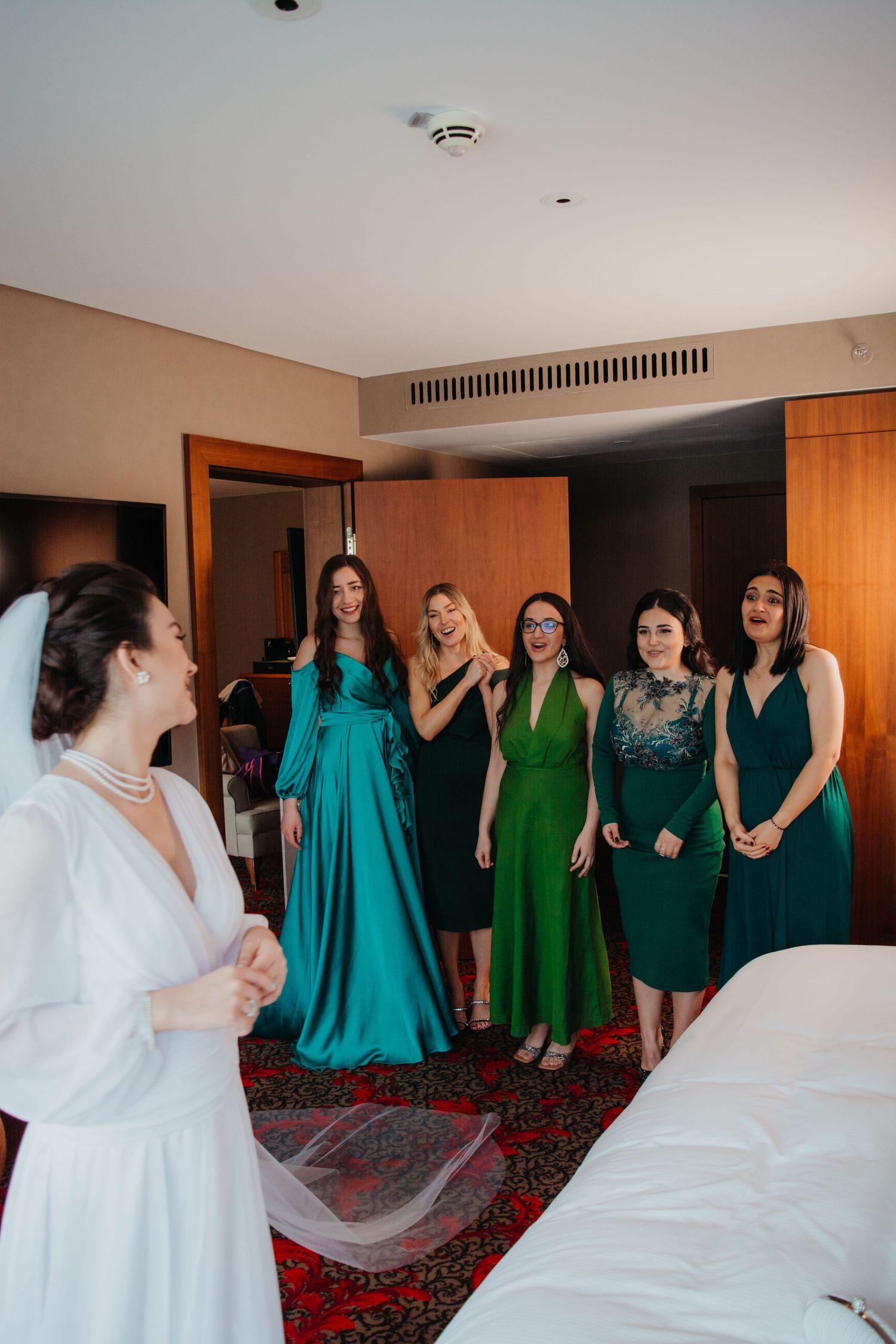 Bride in white gown with veil surprises bridesmaids in green dresses in elegant hotel room.