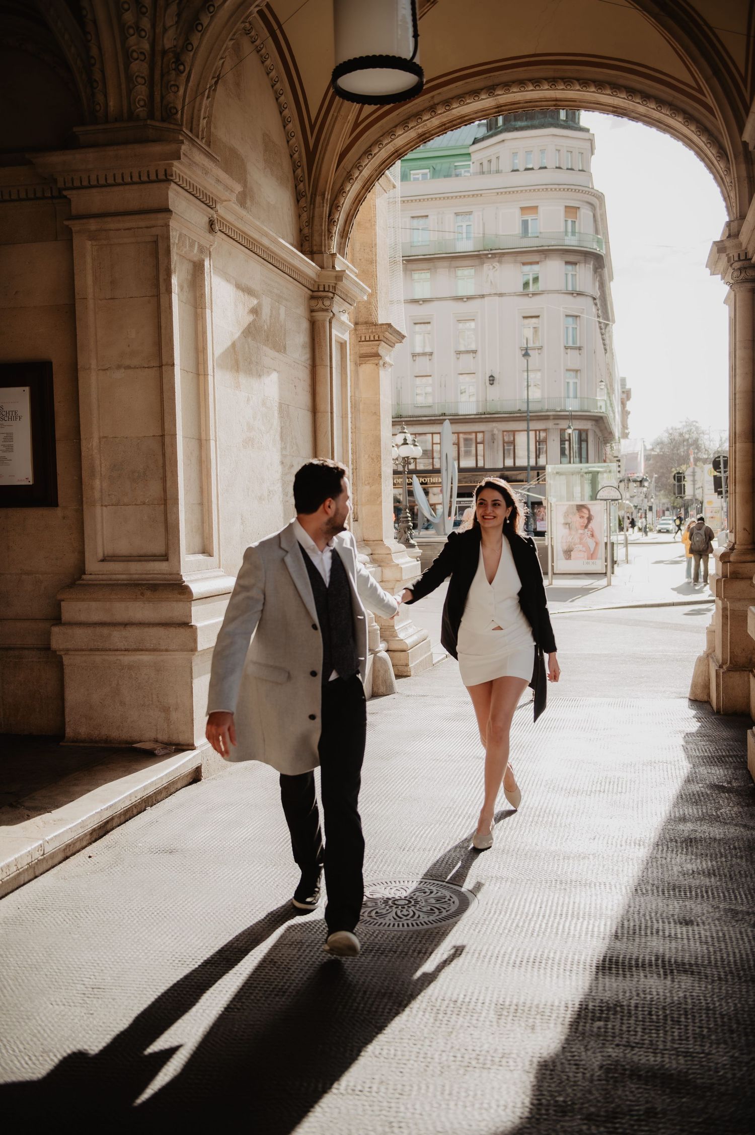 Stylish couple walking hand in hand through a grand European archway in chic black and white outfits.