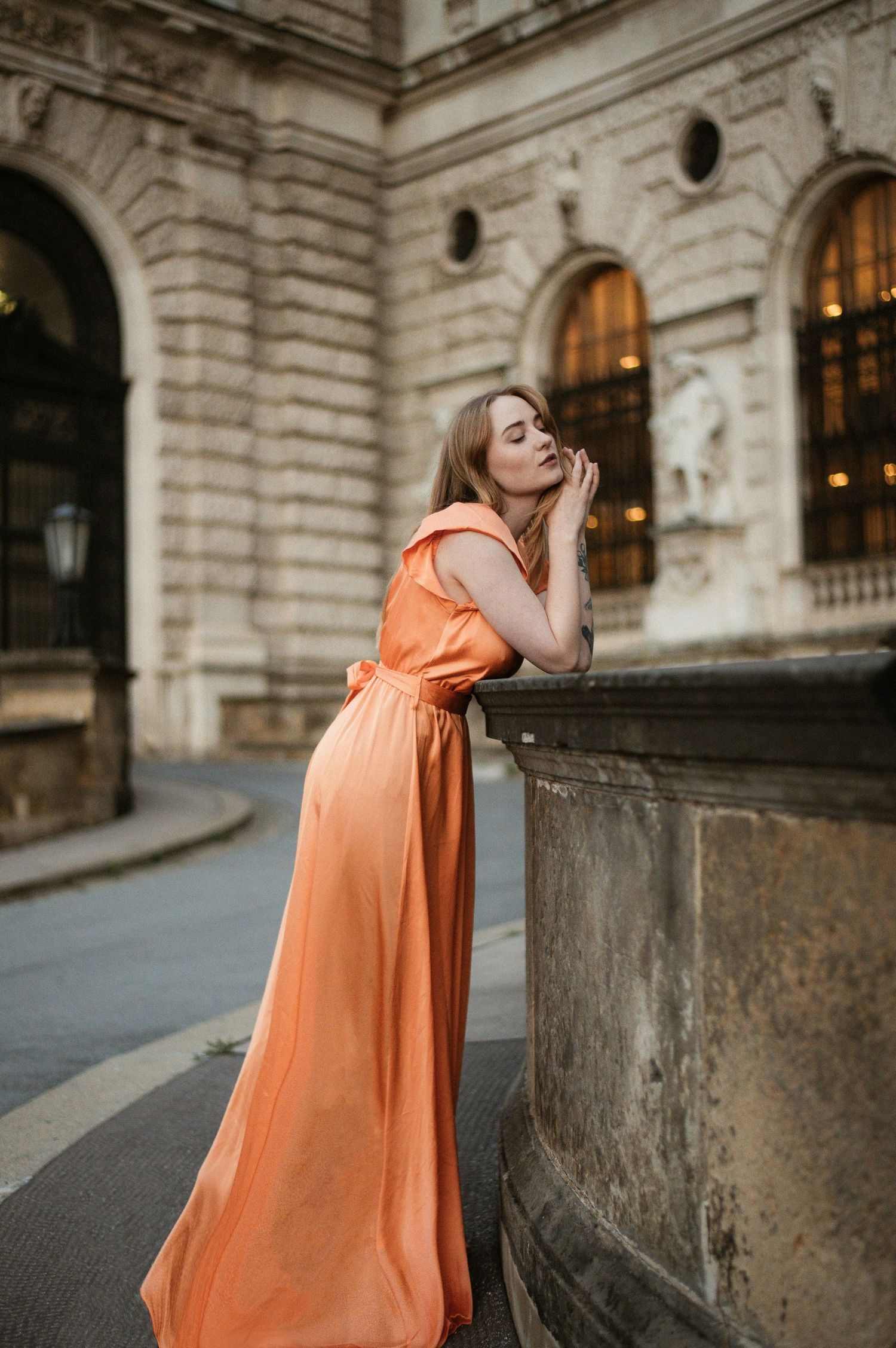 Woman in elegant orange maxi dress leans against stone pillar outside ornate European building at dusk.