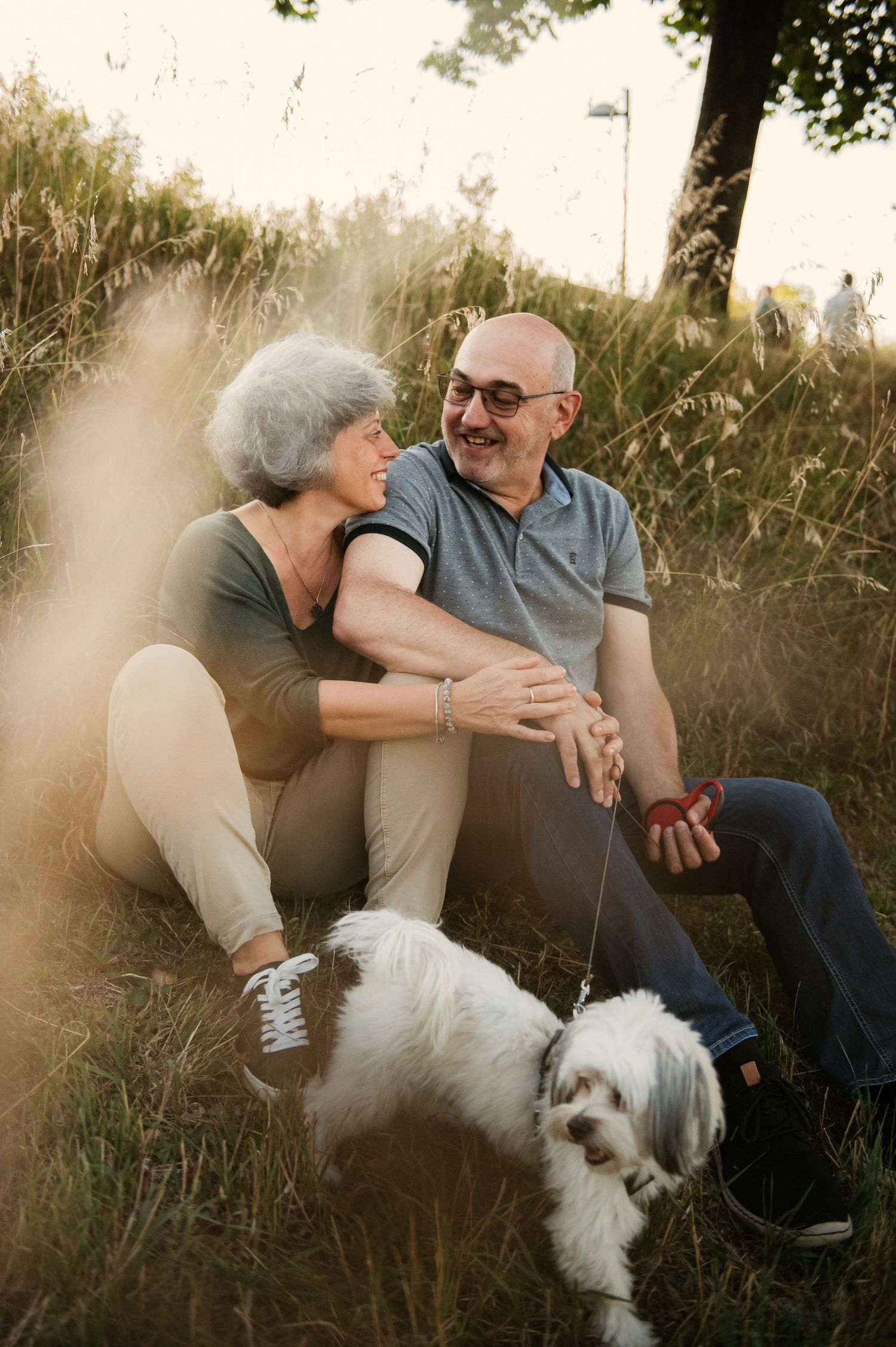 Happy senior couple sitting in grass with small white dog on leash, laughing together outdoors in golden sunlight.