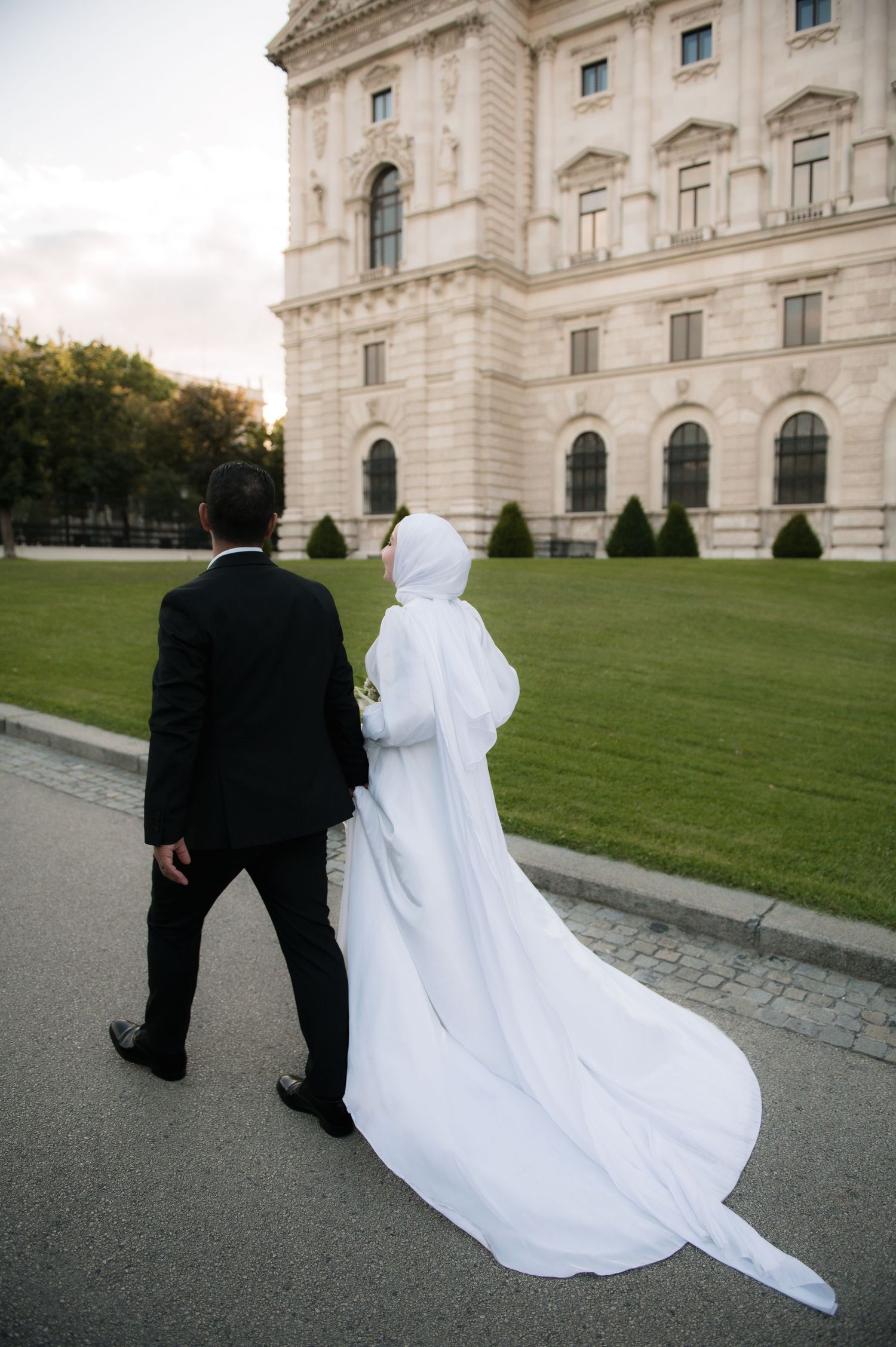 A bride in white hijab and gown walks with groom in black suit near an elegant building with green lawn.