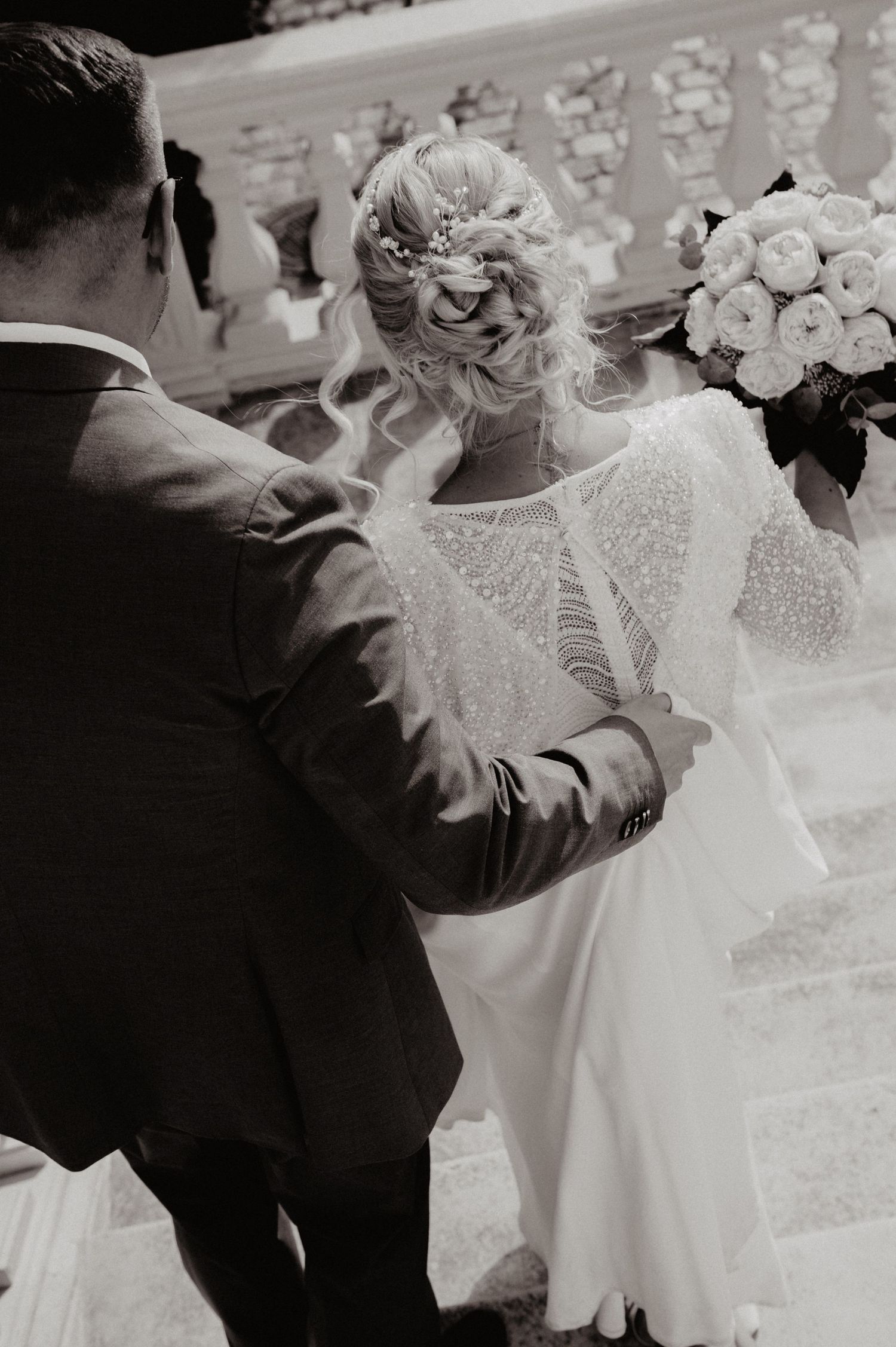 Black and white photo of bride and groom holding hands, bride wearing elegant lace gown with floral updo holding bouquet.