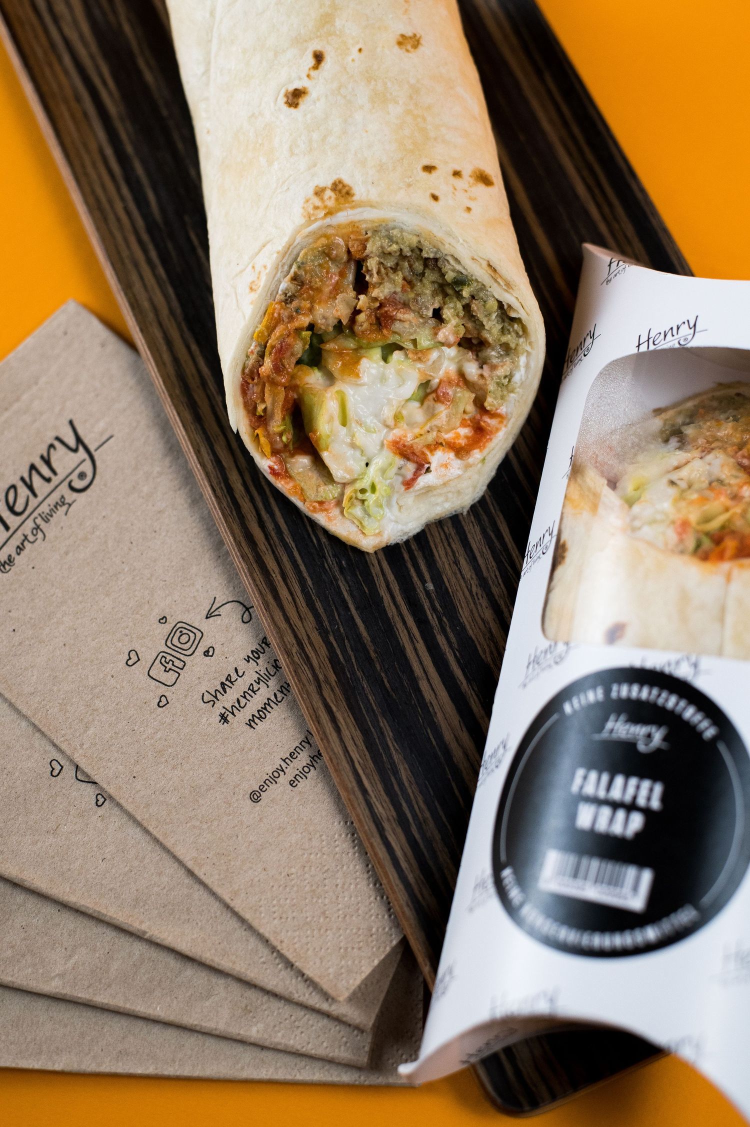 Henry Falafel Wrap on wooden board with branded napkins and packaging on orange background.