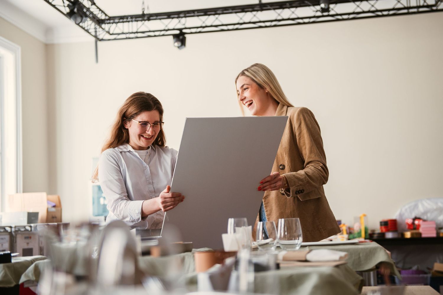 Two women smiling while reviewing a gray board sample in a bright event planning space with tables.