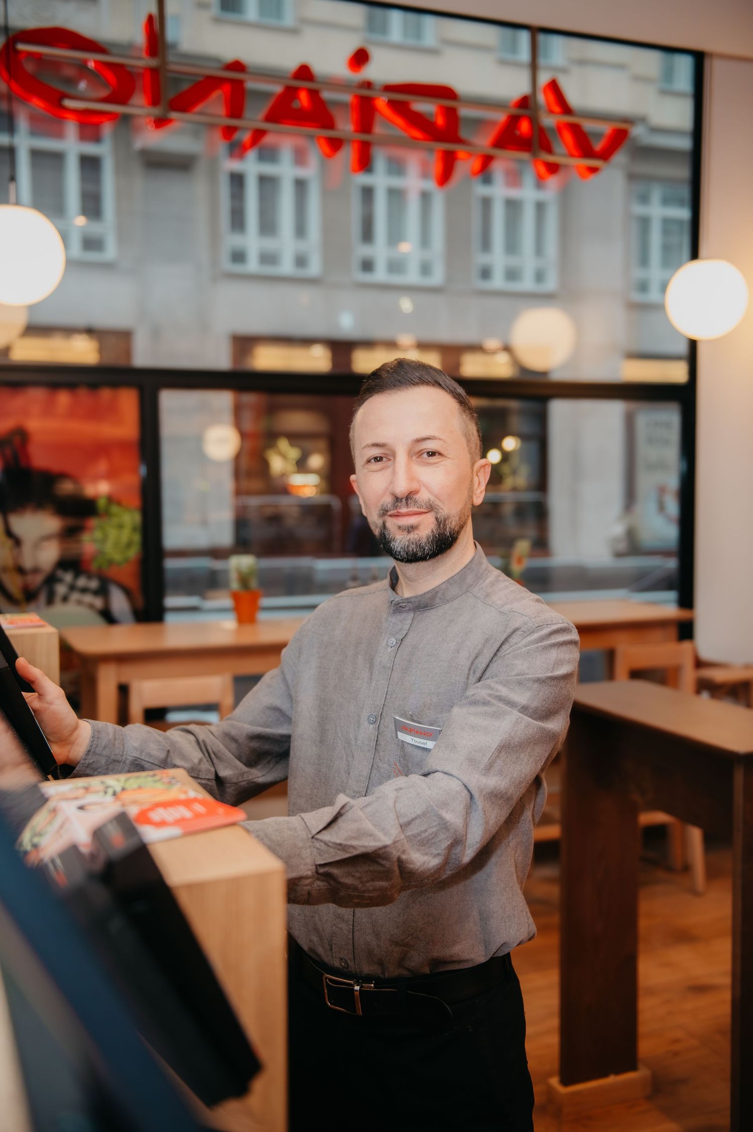 A smiling male restaurant manager in a gray shirt stands at the counter of a modern dining establishment.