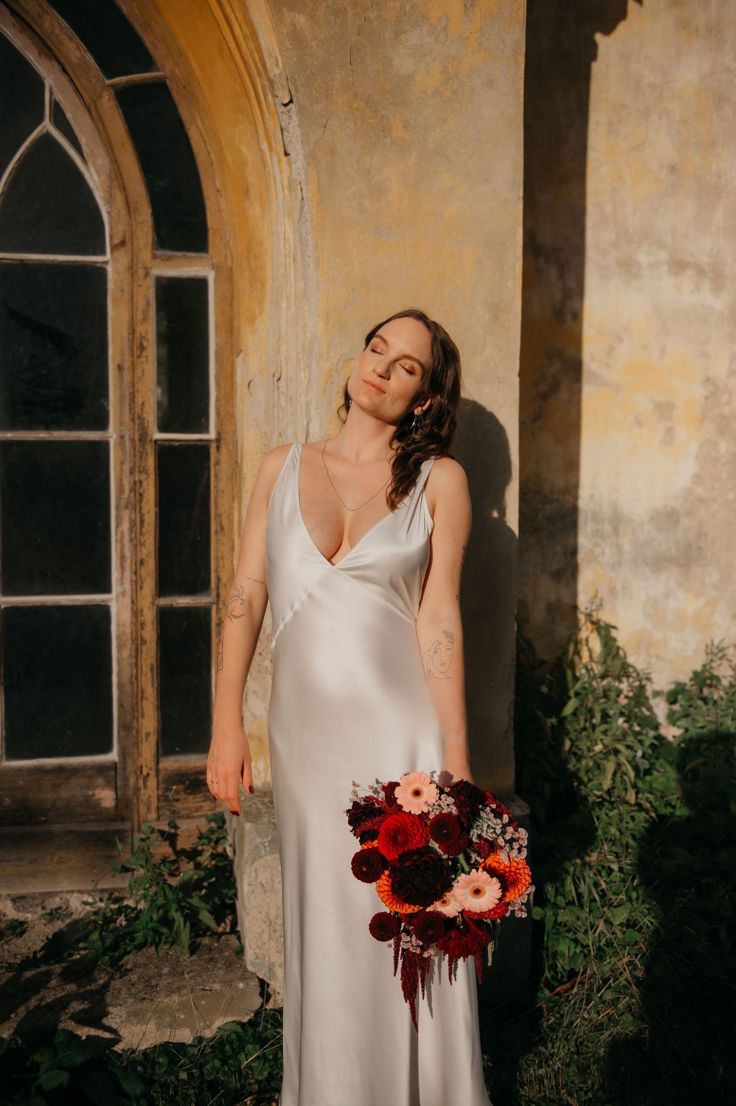 Bride in silver satin gown holds deep red bouquet, posing against rustic yellow wall with arched window.