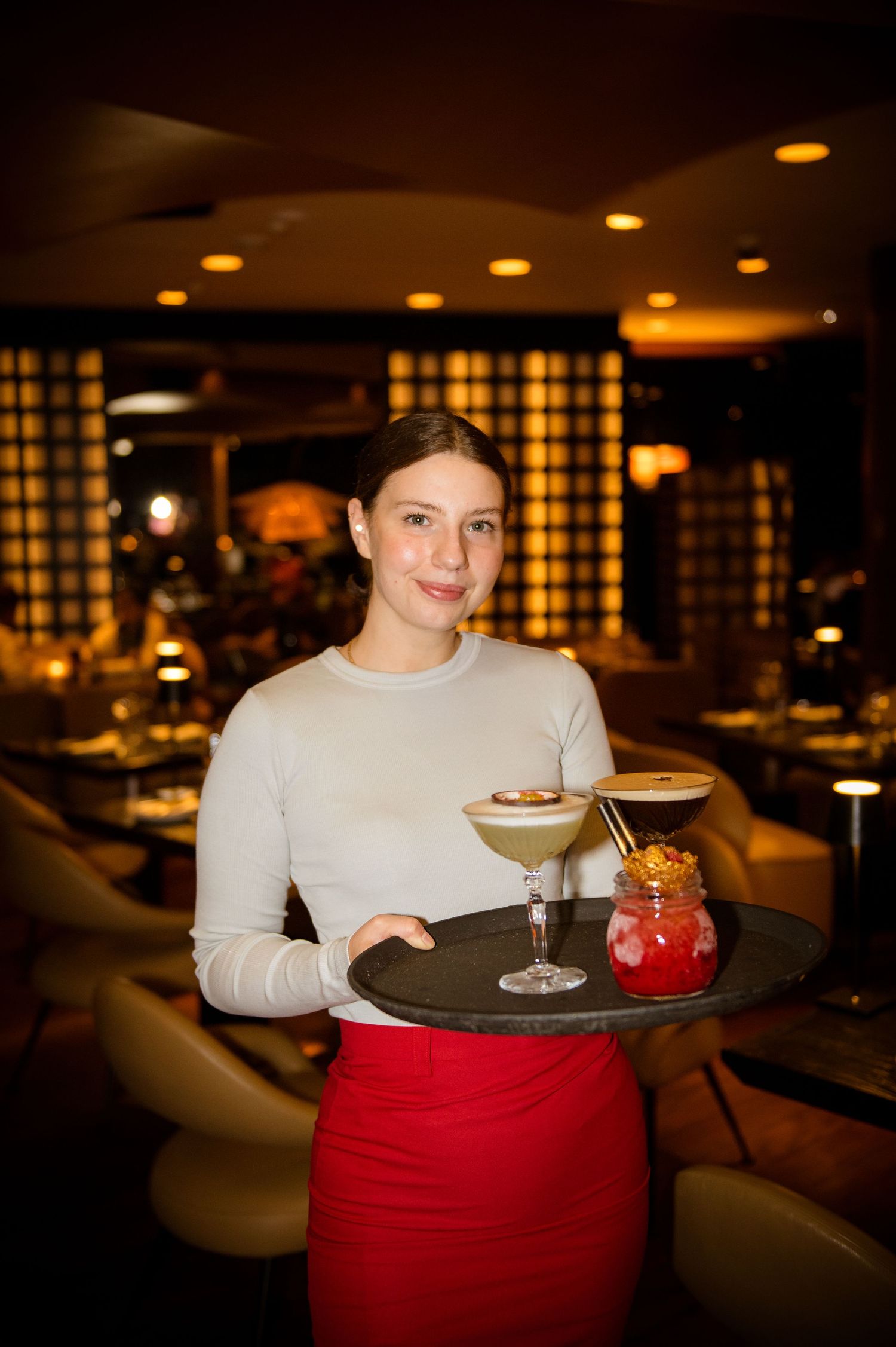 Smiling waitress in red skirt holds tray with cocktails in dimly lit upscale restaurant.