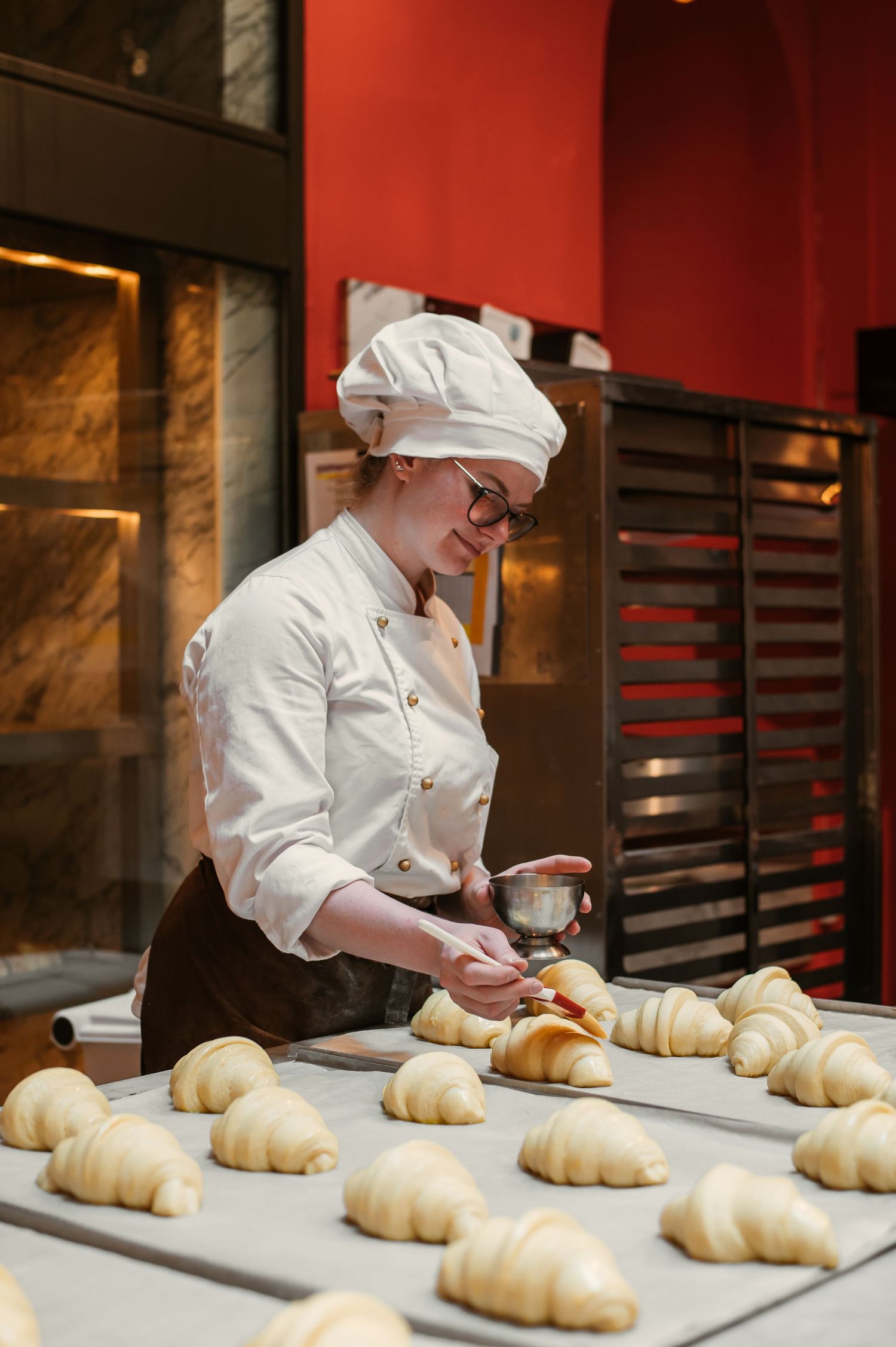 Female baker in white uniform arranging fresh croissants on a baking tray in a professional bakery kitchen.