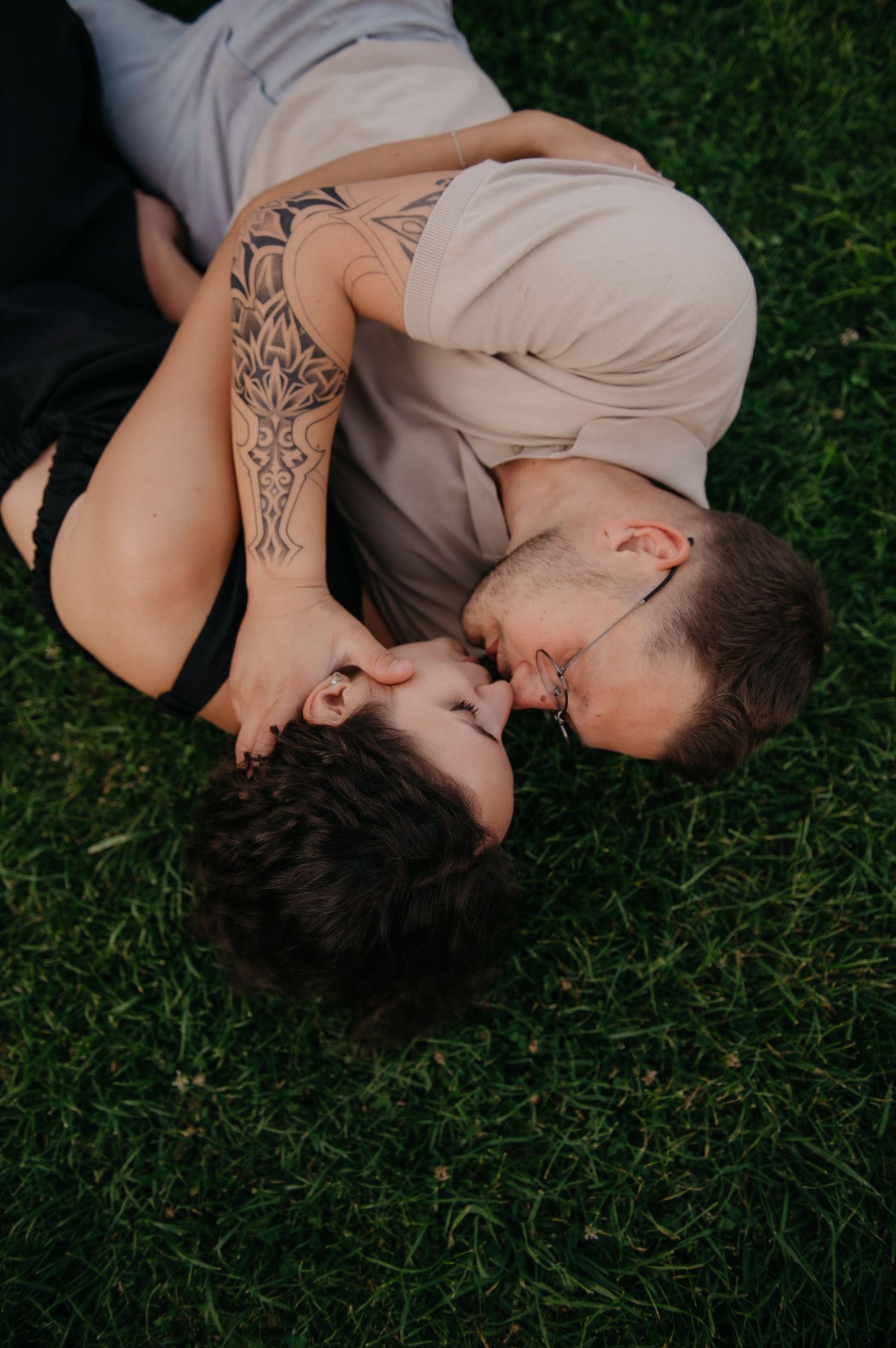 Couple lying on green grass sharing a kiss, one partner with tribal tattoos on arm.