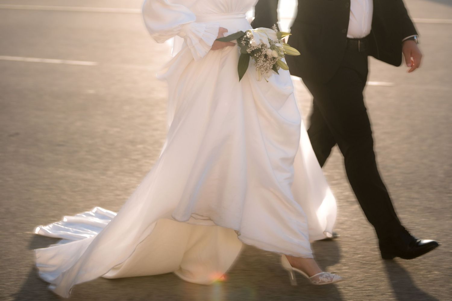 Bride in a flowing white gown with train walking alongside groom in black tuxedo in golden sunlight.