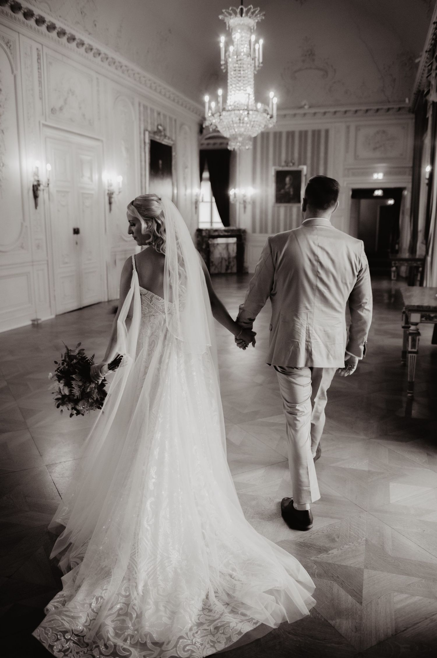 Black and white photo of bride in lace gown and veil holding hands with groom in elegant ballroom.