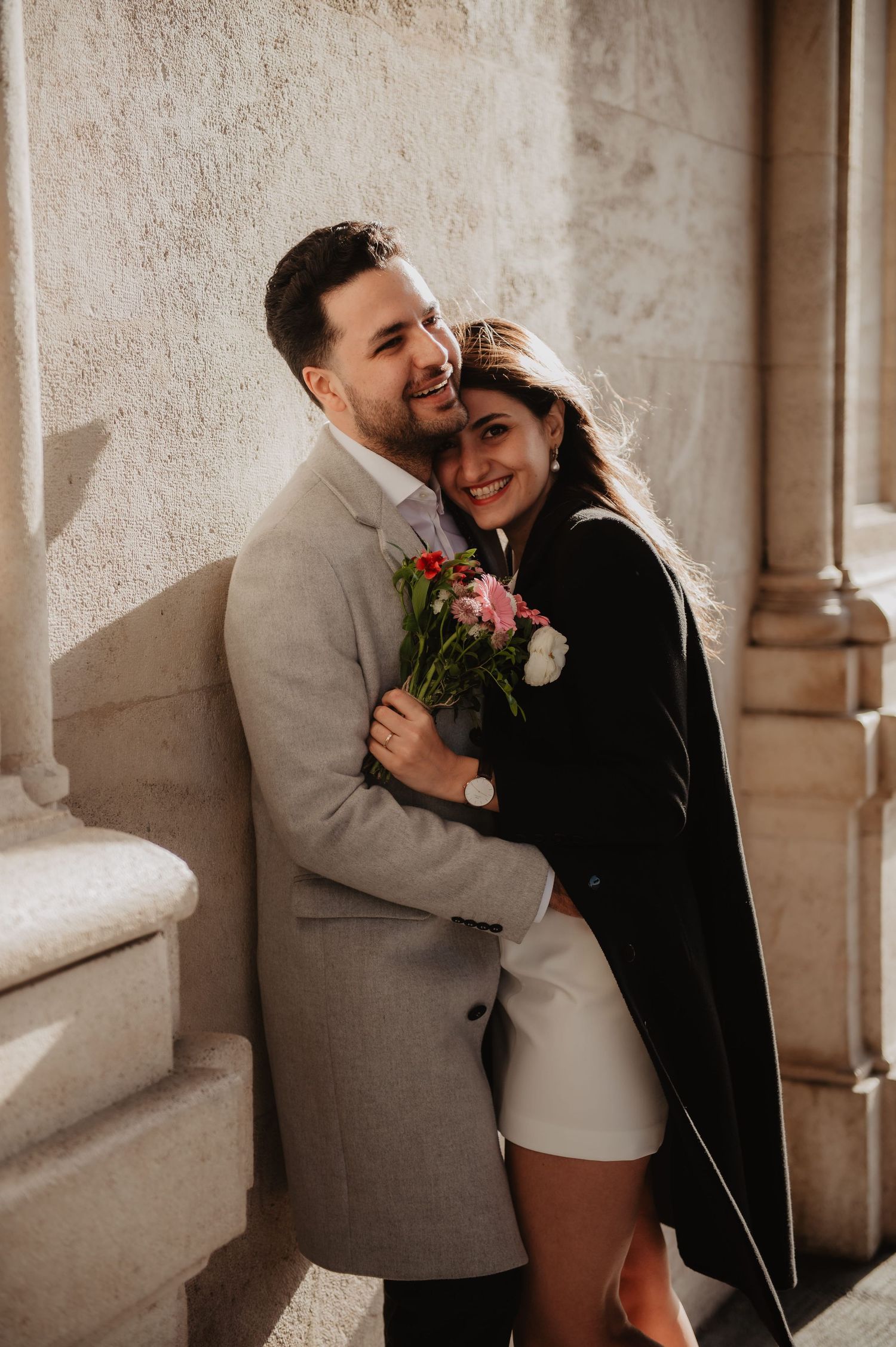 Happy couple embracing against a stone column, woman holding a bouquet of pink and white flowers.
