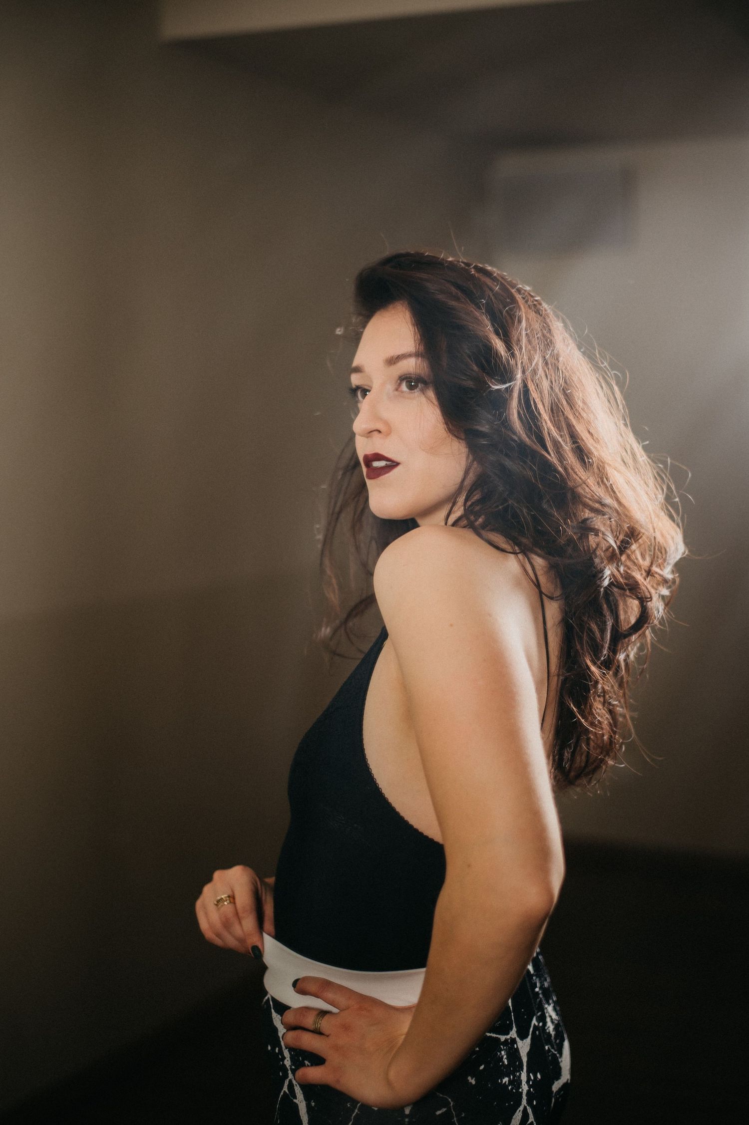 Brunette woman in a black backless dress poses dramatically in a smoky dimly lit studio setting.