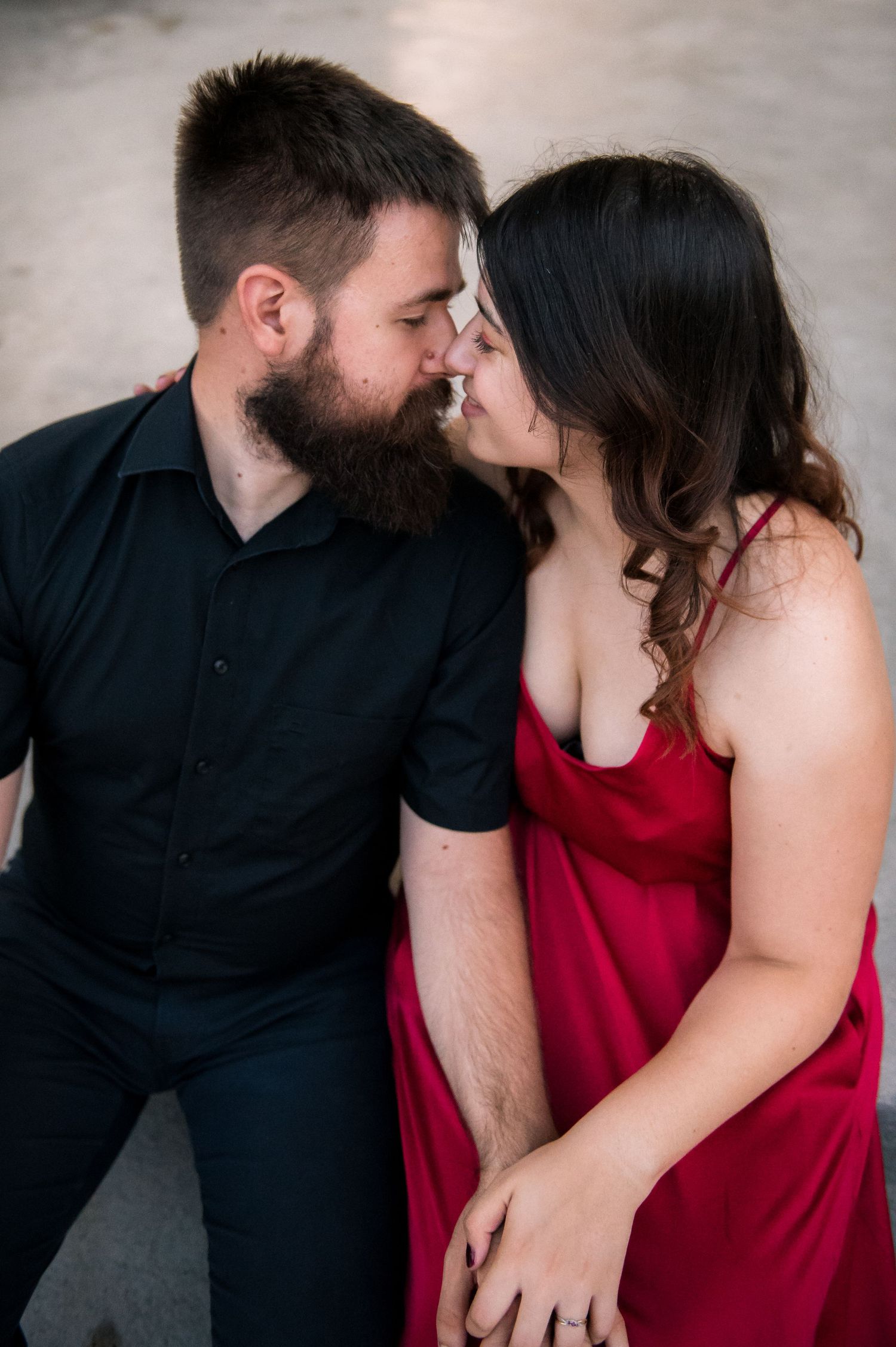 A bearded man in black shirt and woman in red dress share a romantic almost-kiss moment while holding hands.