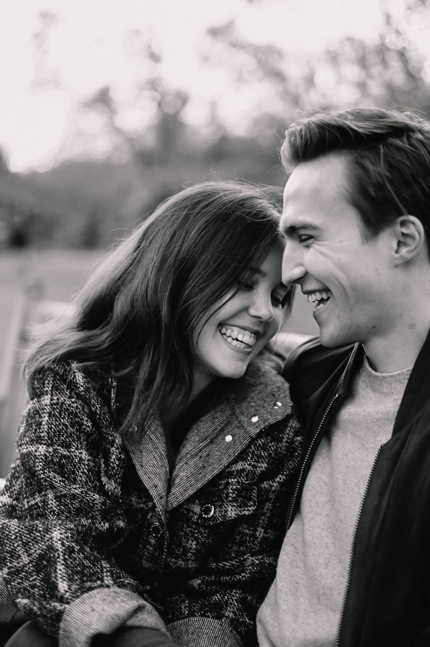 Black and white photo of a smiling couple leaning close together outdoors in autumn, wearing cozy jackets.