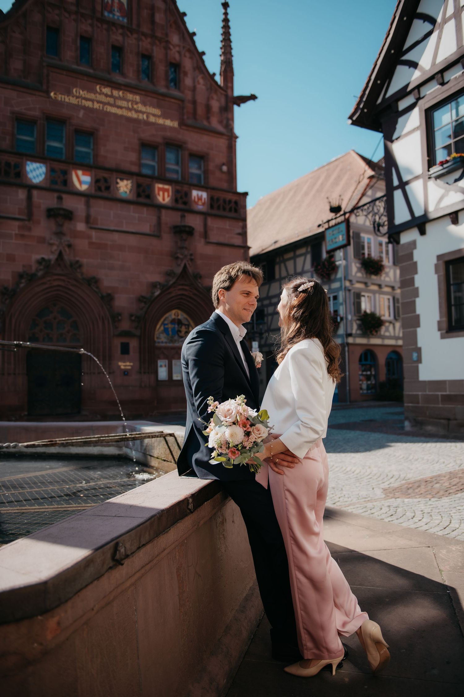 Couple in wedding attire embracing by a fountain in a European town square with historic buildings.