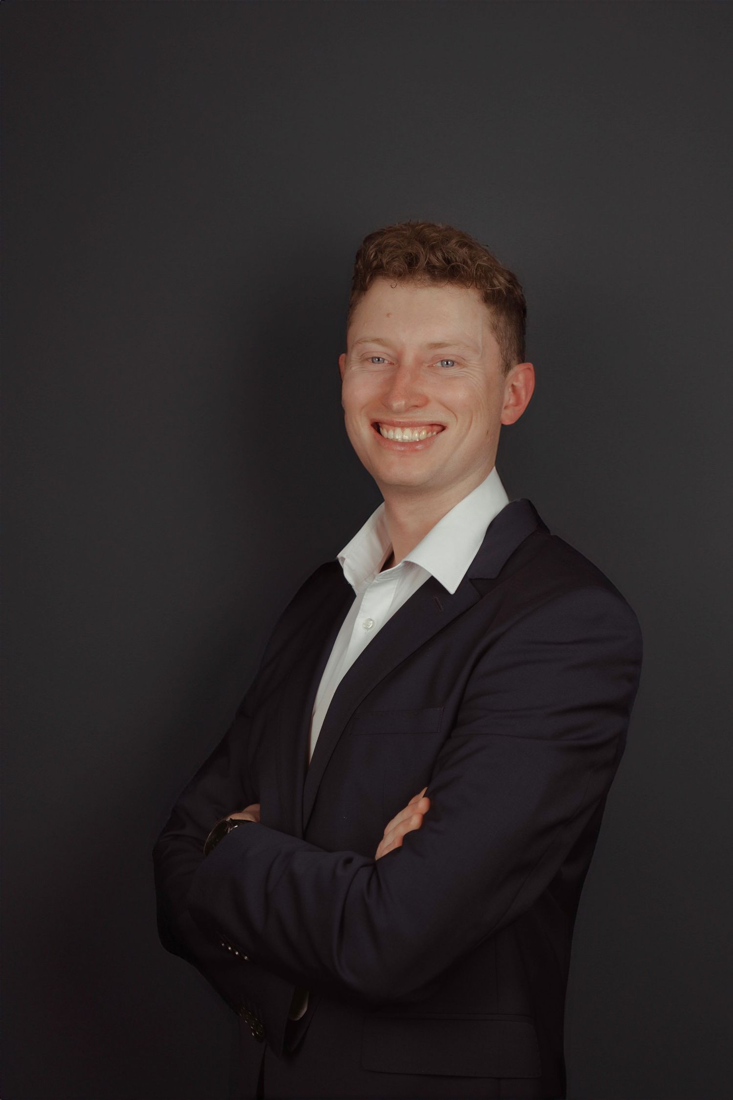 Young professional man in dark suit smiling with arms crossed against gray background.