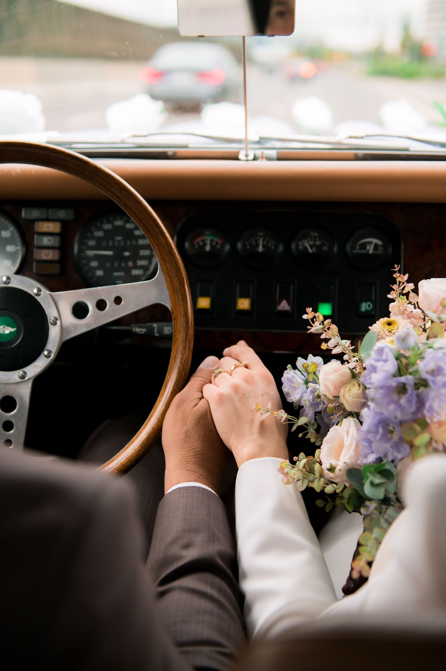 Newlyweds holding hands in a vintage Jaguar, bride holding pastel floral bouquet, wedding rings visible.