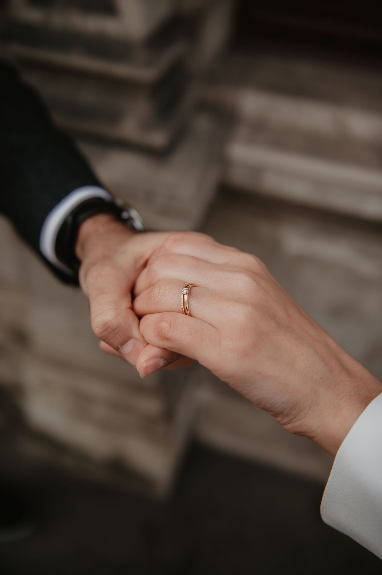 Close-up of two hands holding, one wearing a gold engagement ring, with a blurred stone background.