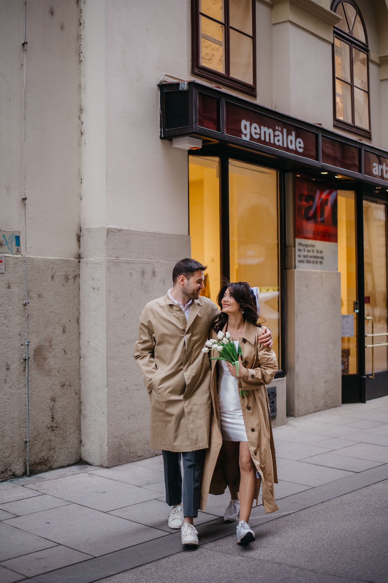 Couple in matching beige trench coats walking together outside a gemalde art gallery, woman holding white tulips.