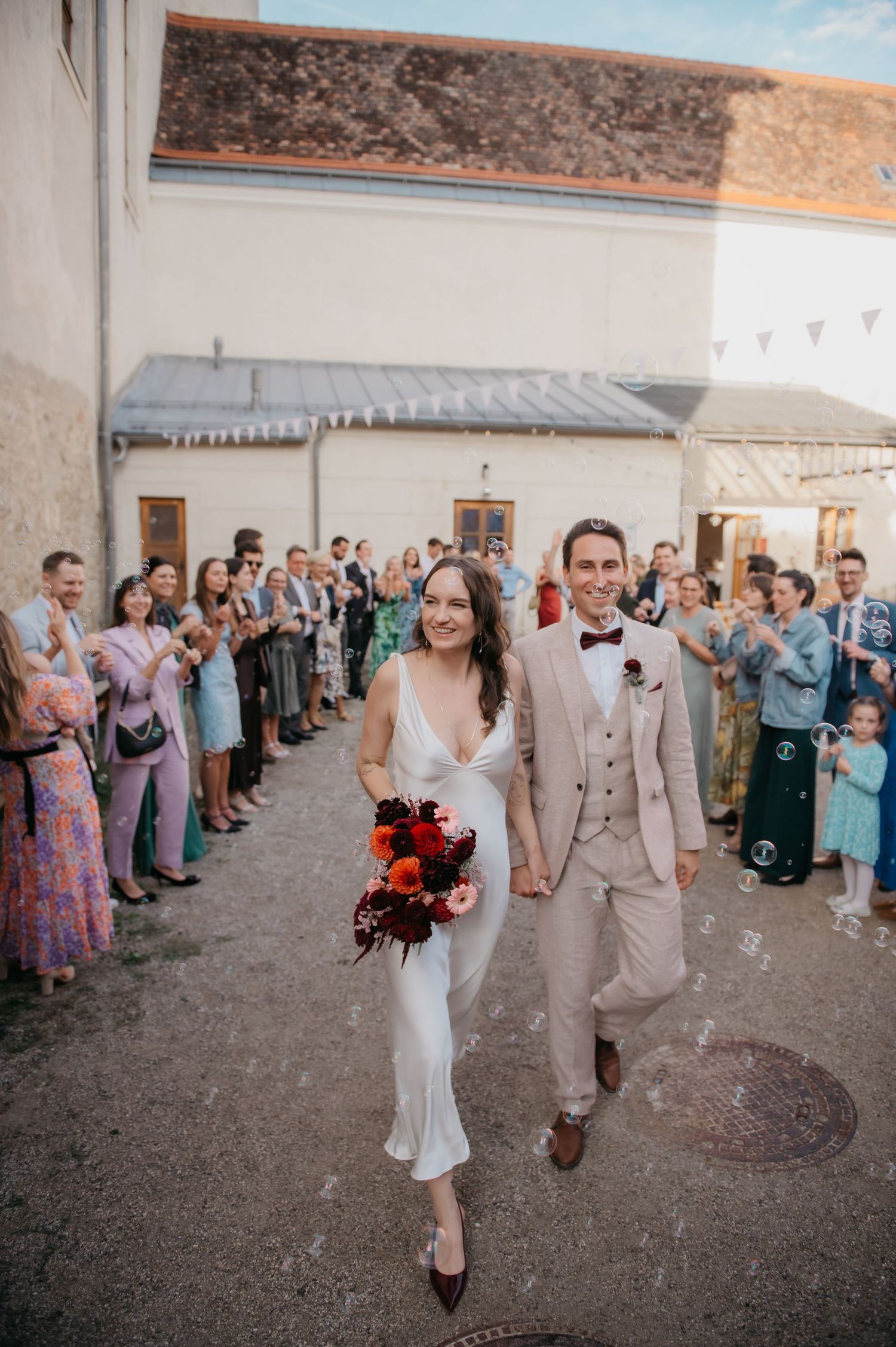 Newlywed couple walks hand-in-hand through cheering wedding guests in a charming outdoor courtyard ceremony exit.