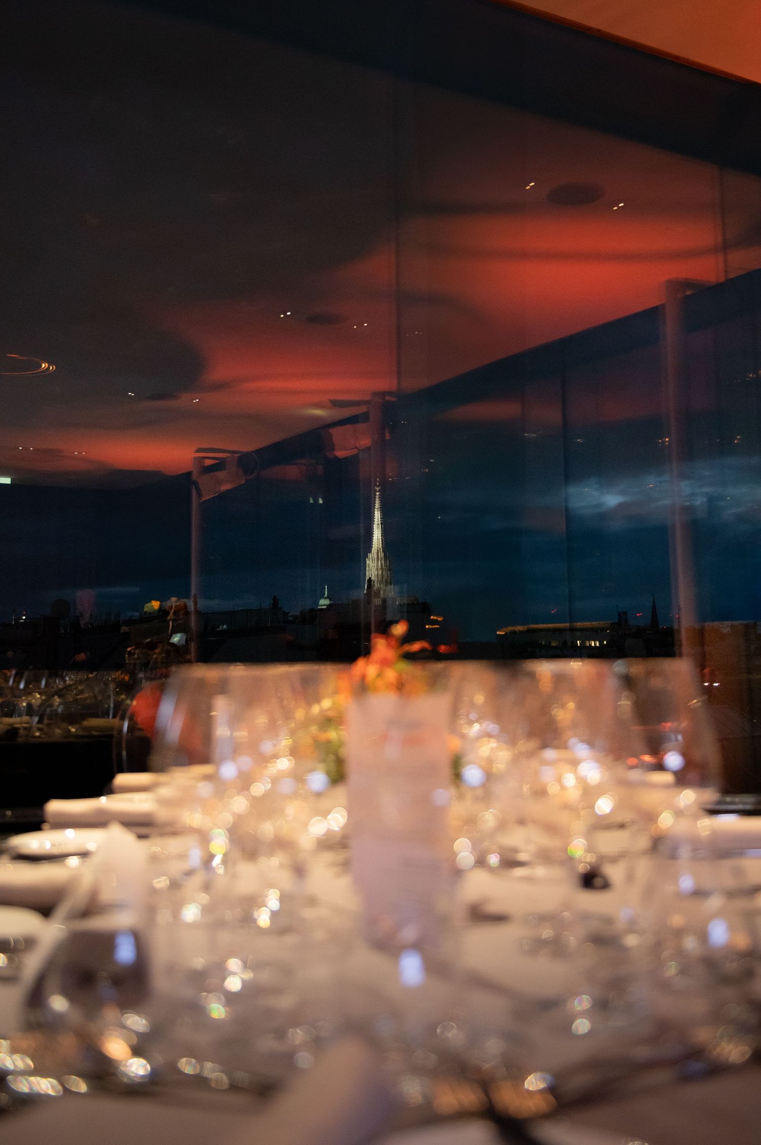 Elegant rooftop dining table set with crystal glasses at dusk, city skyline and church spire visible through floor-to-ceiling windows.
