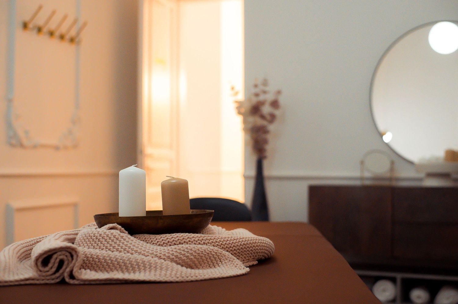 Serene spa massage table with white candles on wooden tray, folded towel, round mirror, and dried flowers in background.