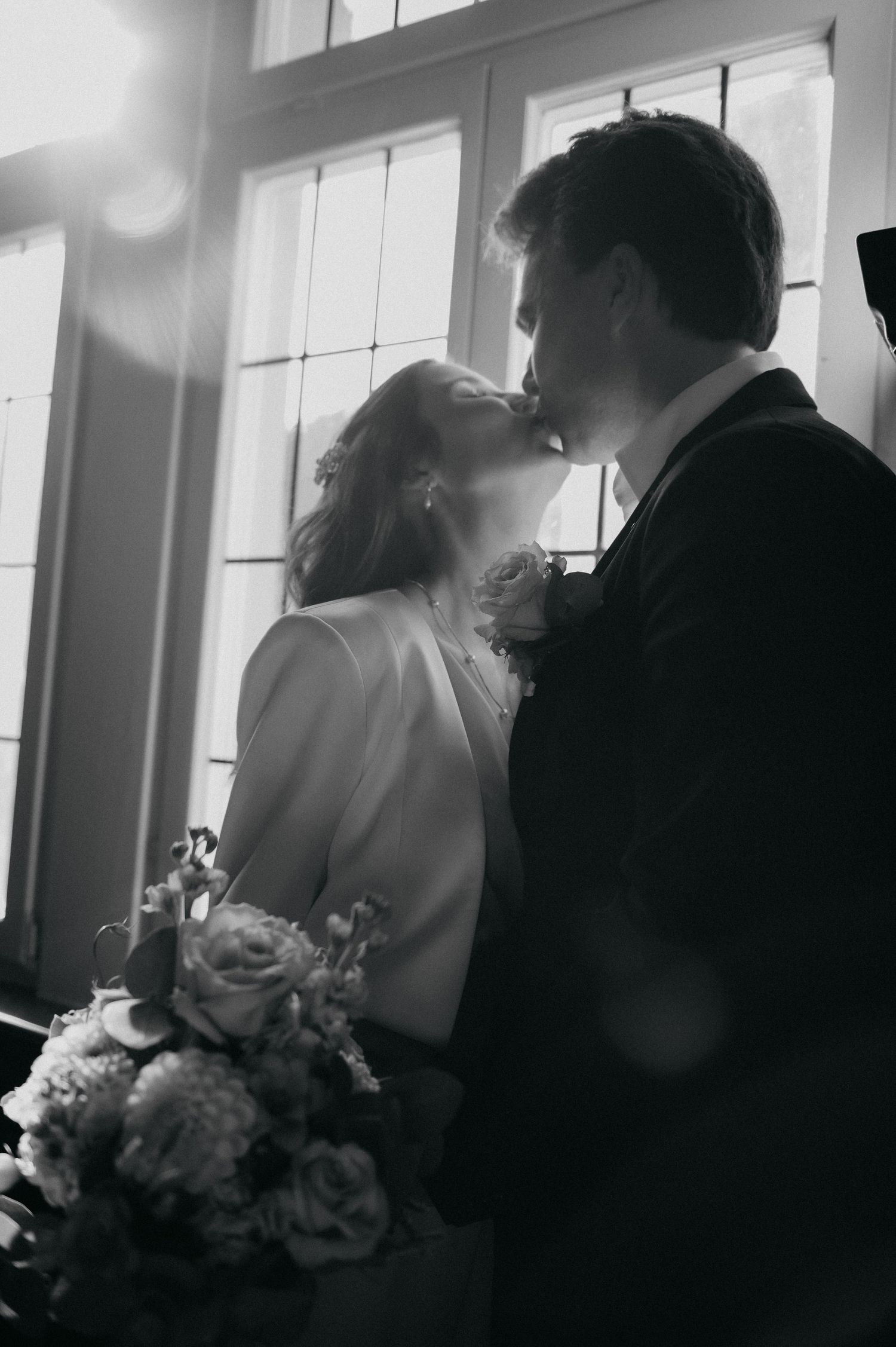 Black and white photo of a bride and groom sharing a kiss, bride holding a floral bouquet, backlit by windows.