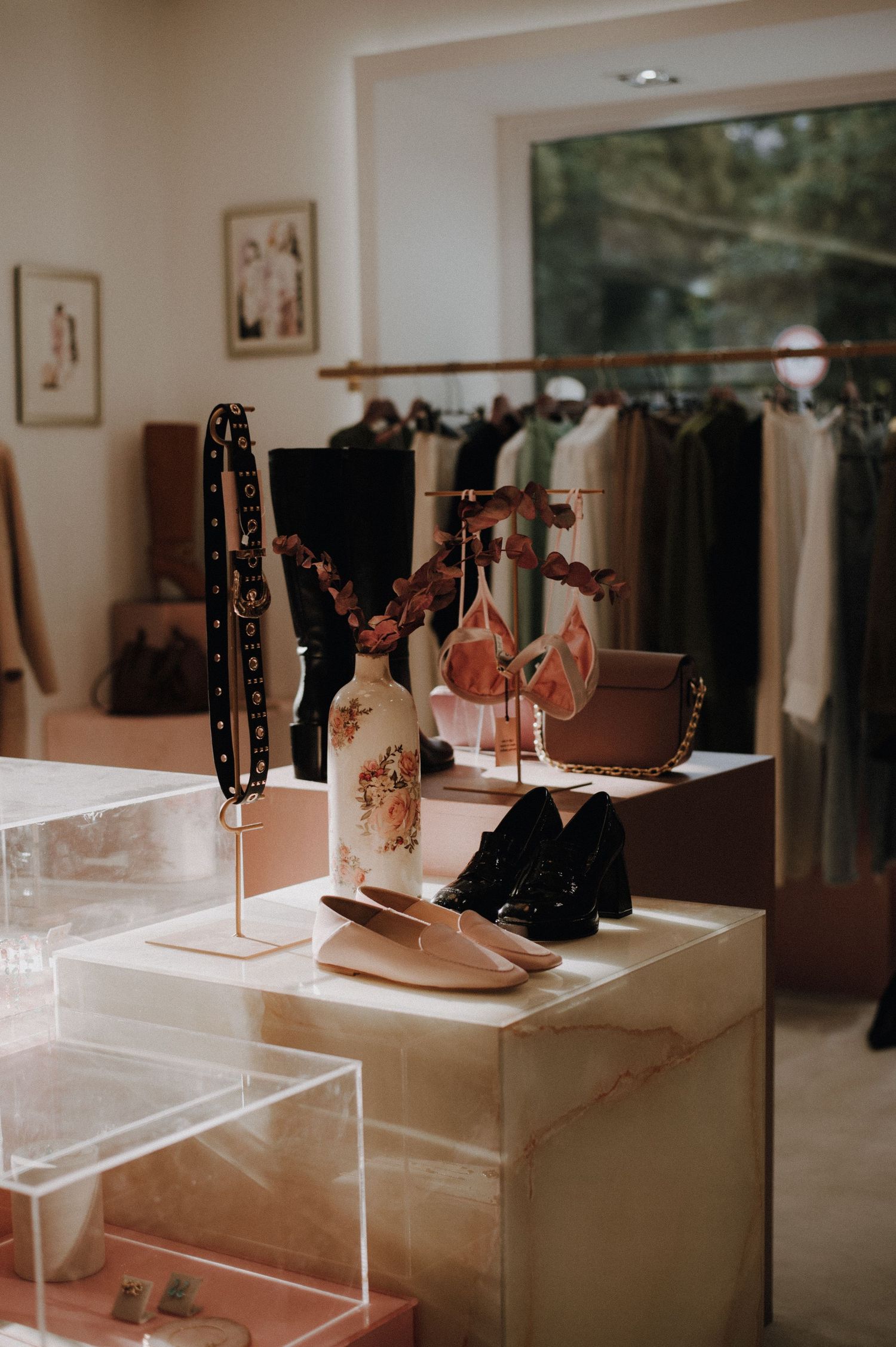 Elegant boutique display with shoes, handbags, and accessories on a marble pedestal with clothing rack in background.