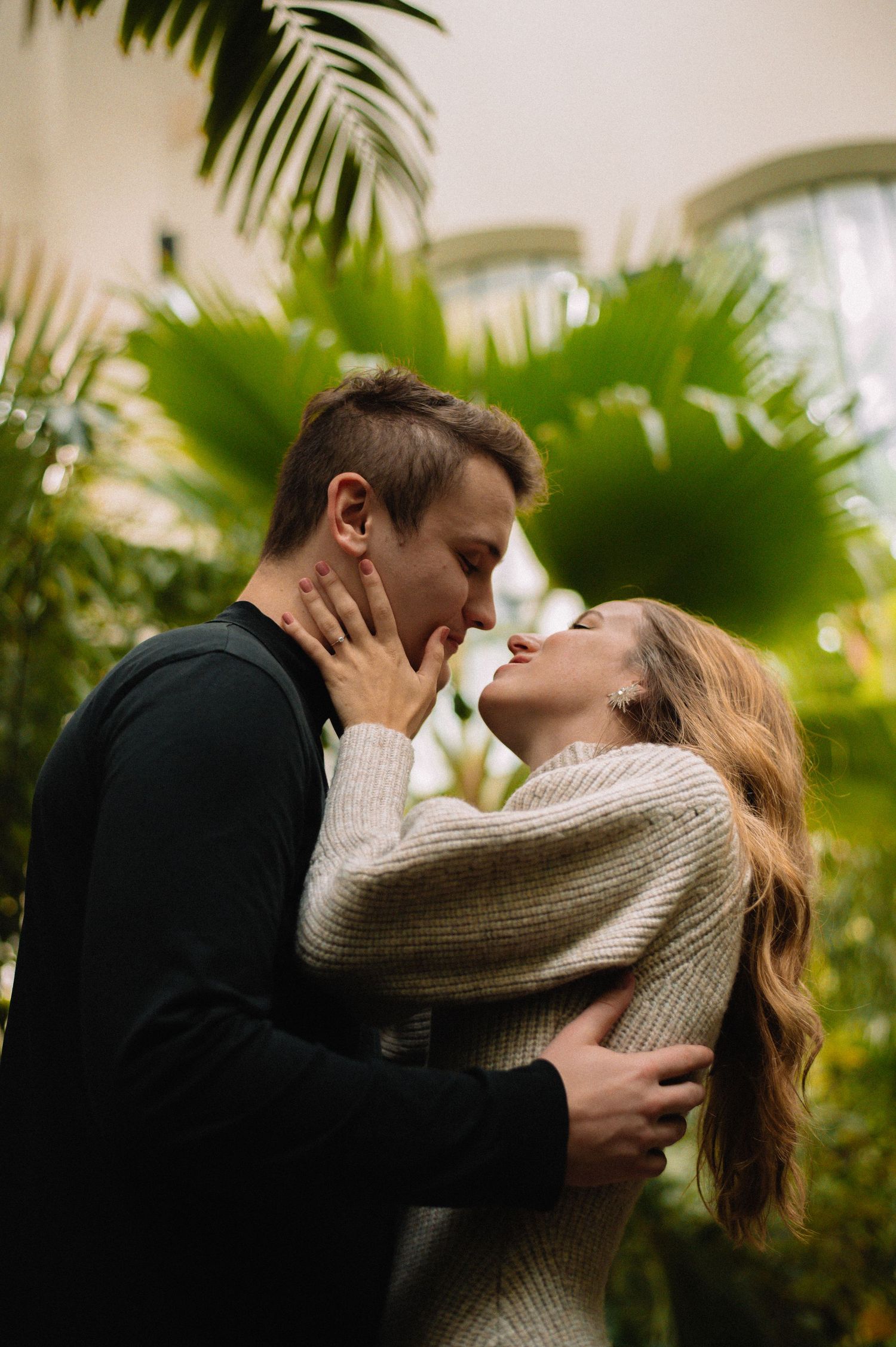 A romantic couple sharing an intimate moment about to kiss surrounded by lush tropical green palm trees indoors.