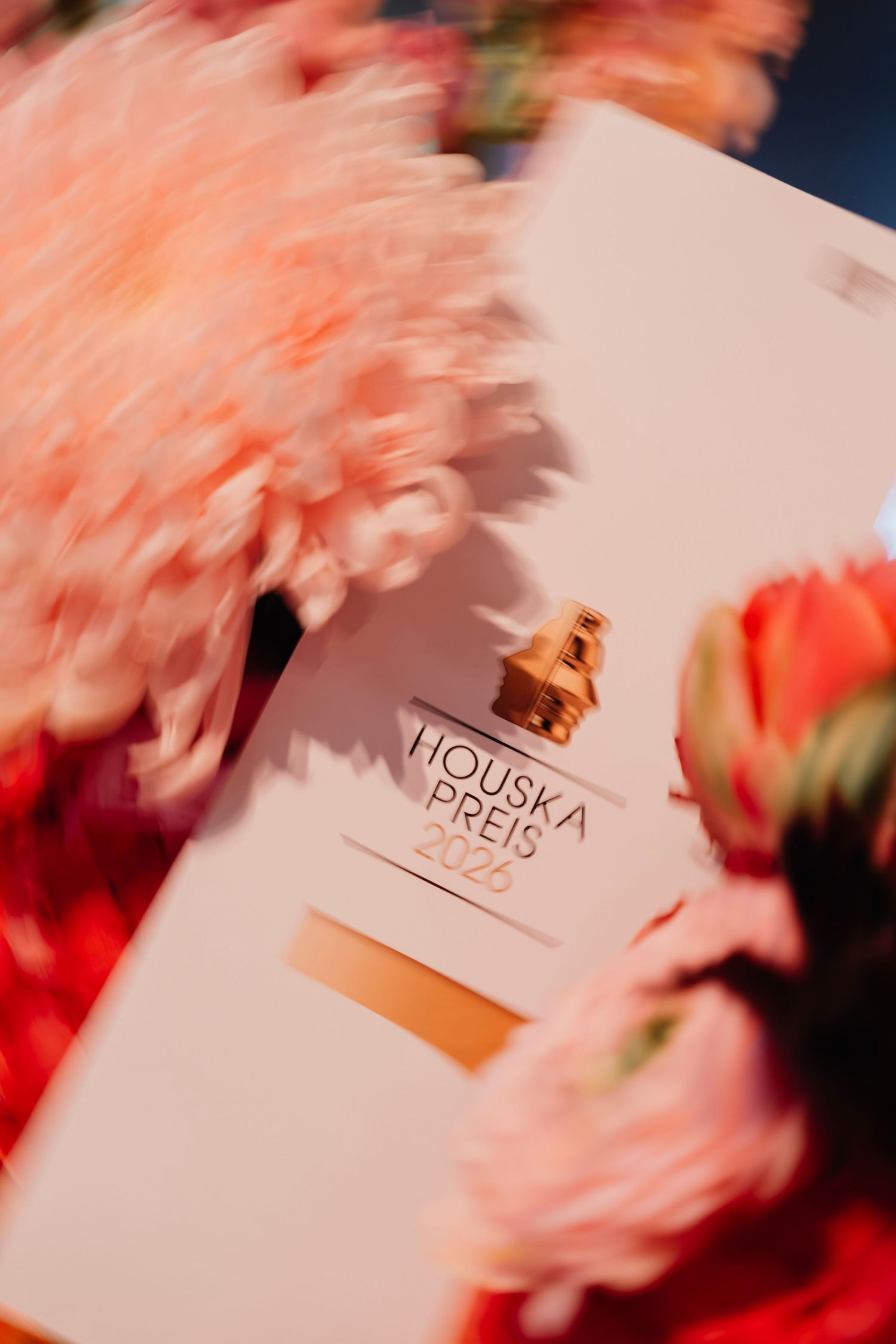 Houska Preis 2023 award booklet with a gold perfume bottle, surrounded by soft pink and red flowers.