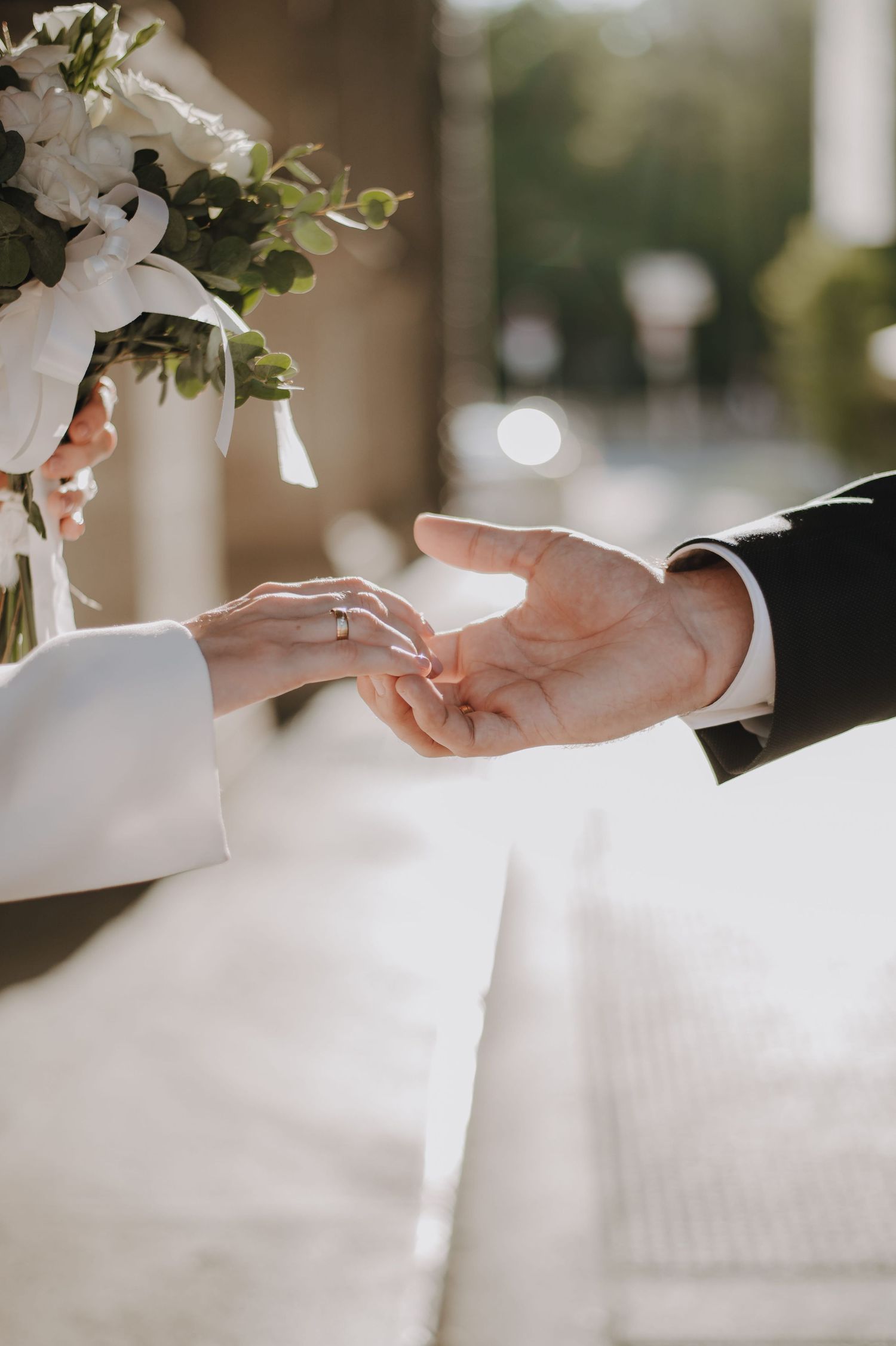 Close-up of bride placing wedding ring on groom's hand outdoors, with white floral bouquet visible in soft light.