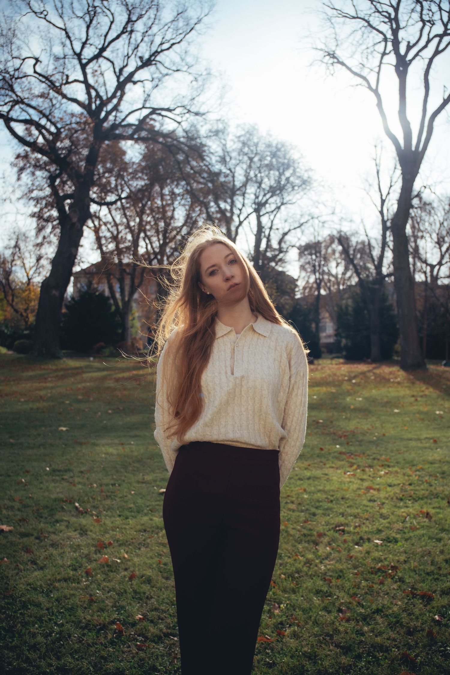 Young woman with long hair in cream sweater and black skirt standing in autumn park with bare trees.
