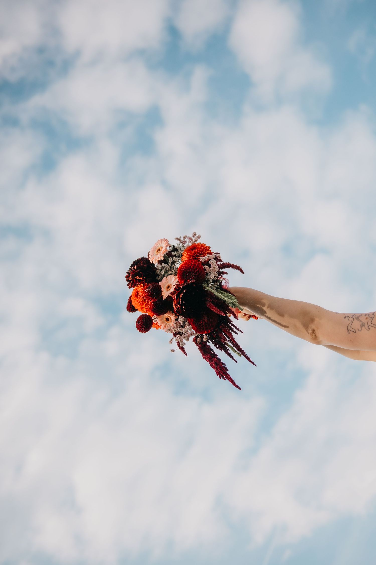 A tattooed hand holds a vibrant red and burgundy flower bouquet against a bright blue cloudy sky.