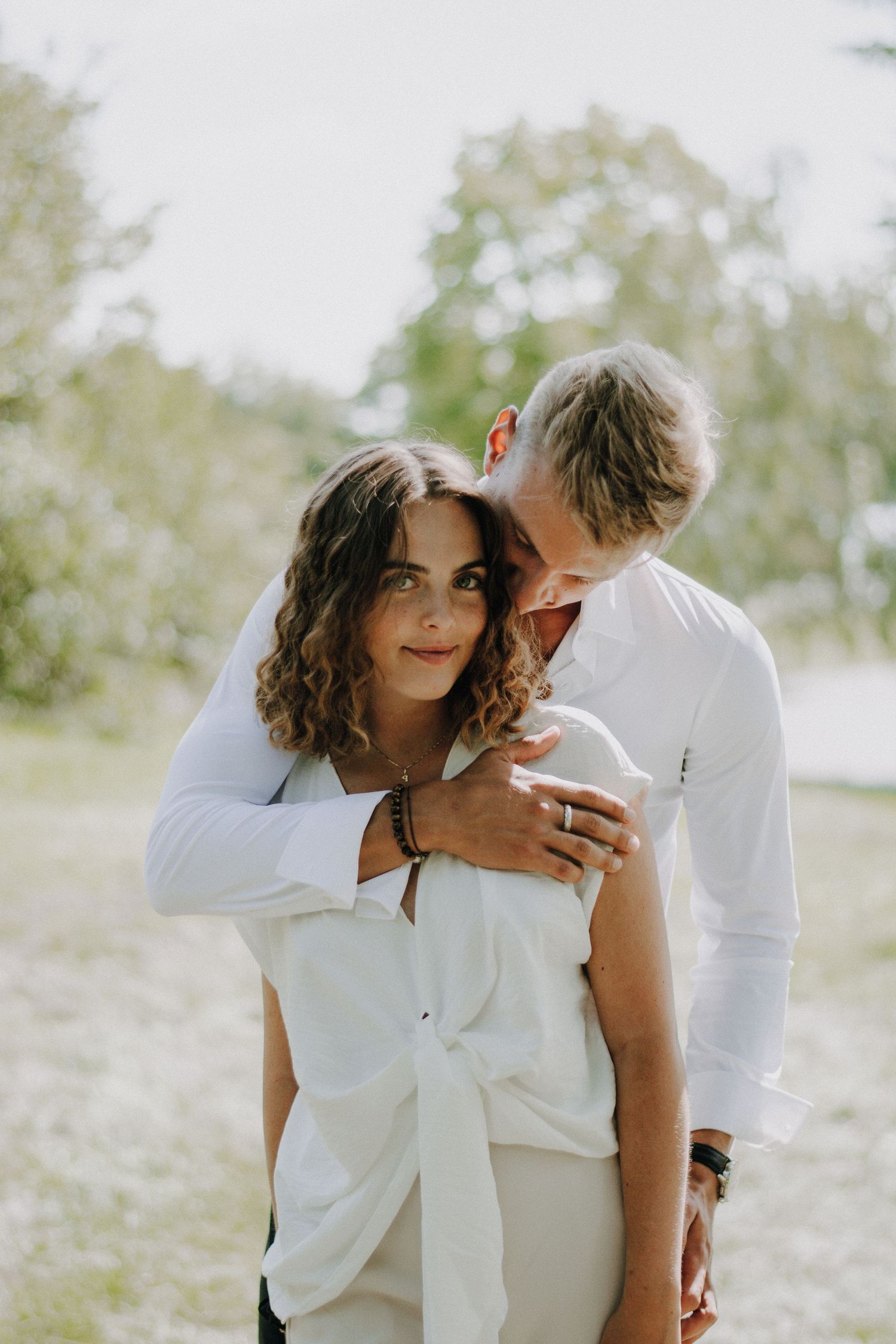 A couple in white outfits embraces outdoors, man nuzzling woman's neck while she smiles softly in a lush green setting.