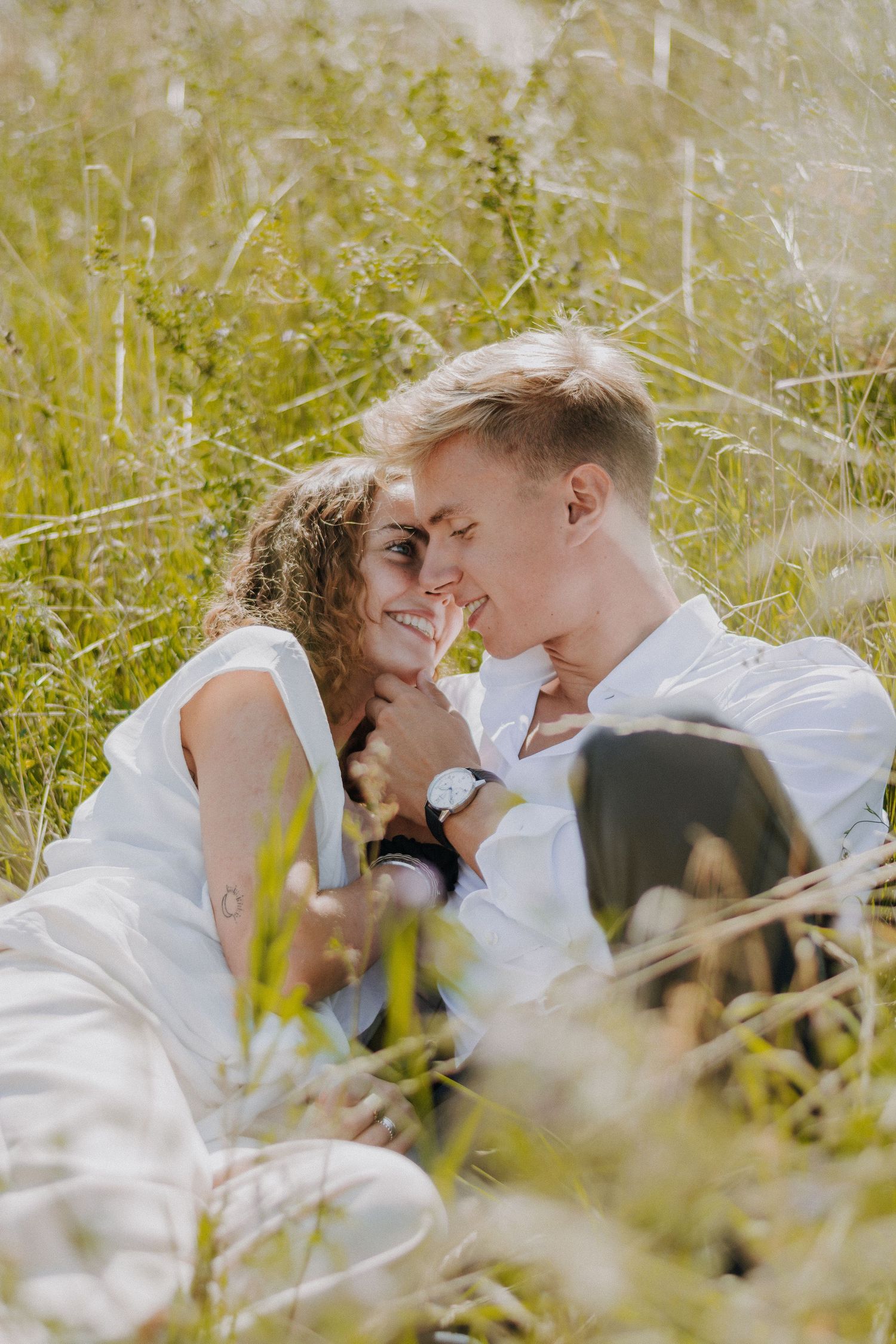 A smiling couple sits together in a lush green field, gazing at each other in a romantic outdoor setting.