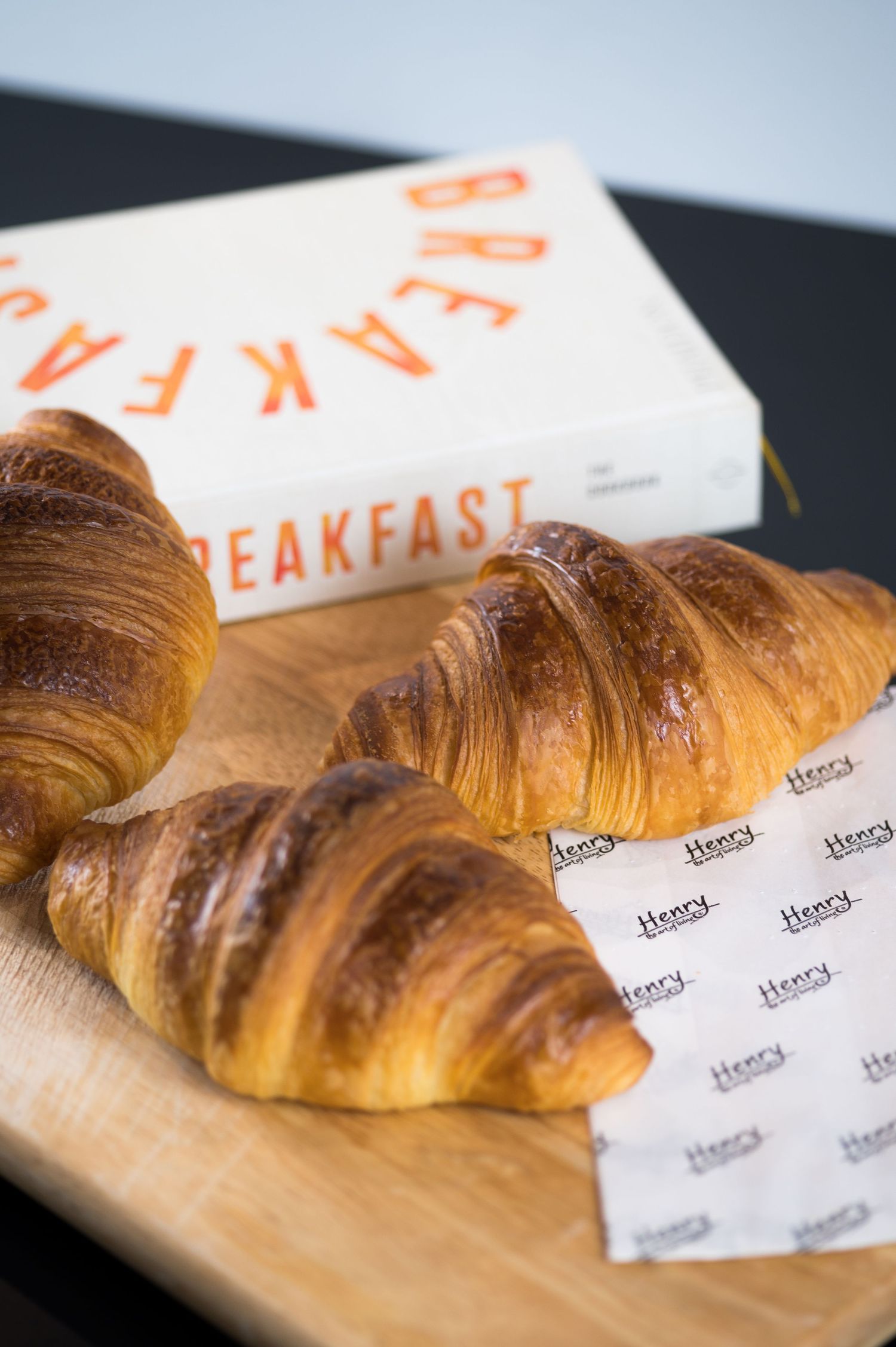 Two golden croissants on a wooden board with Henry-branded paper and a Breakfast box in the background.