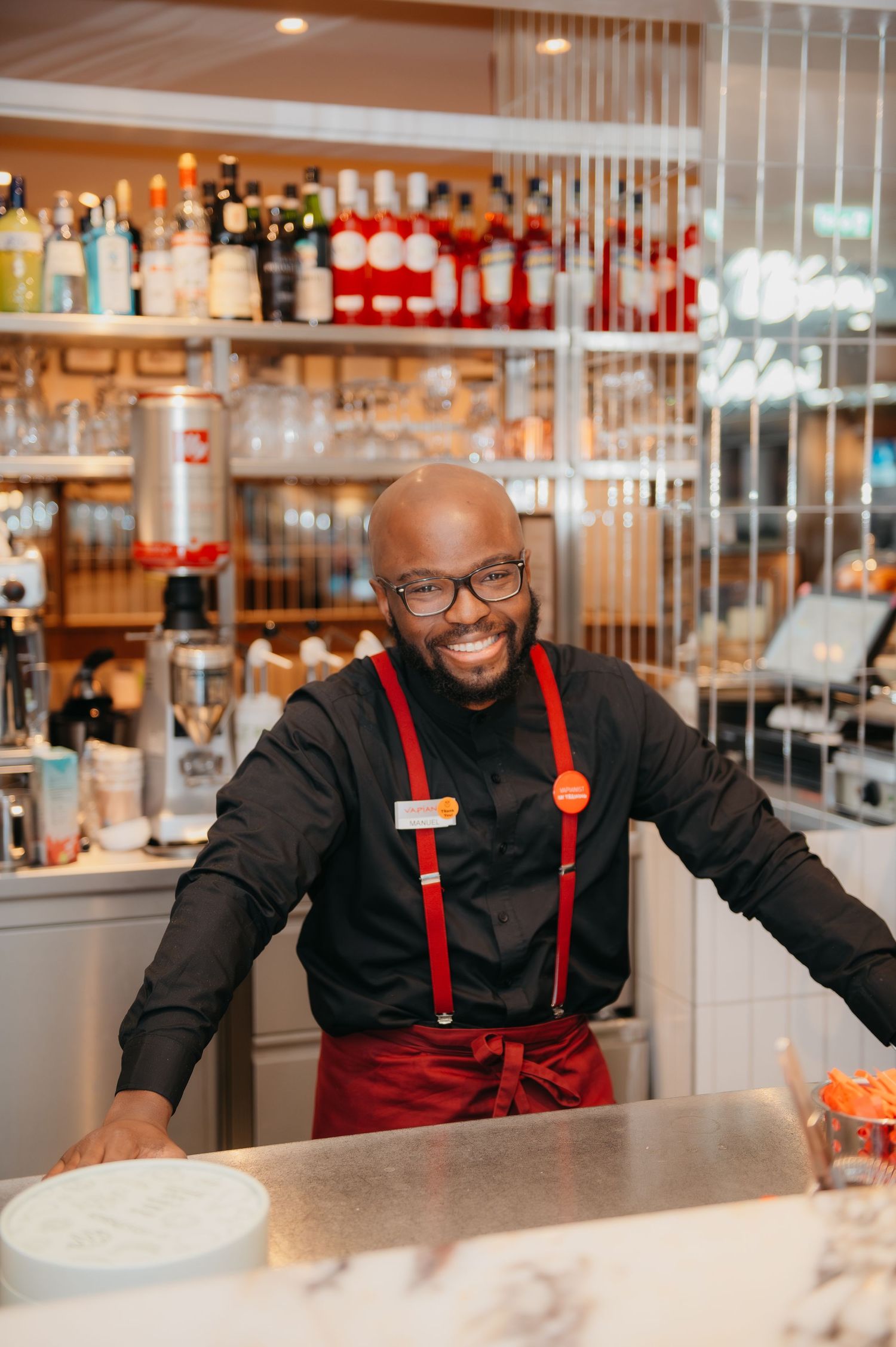 Smiling bartender in black shirt and red suspenders stands behind bar with liquor bottles on shelves.